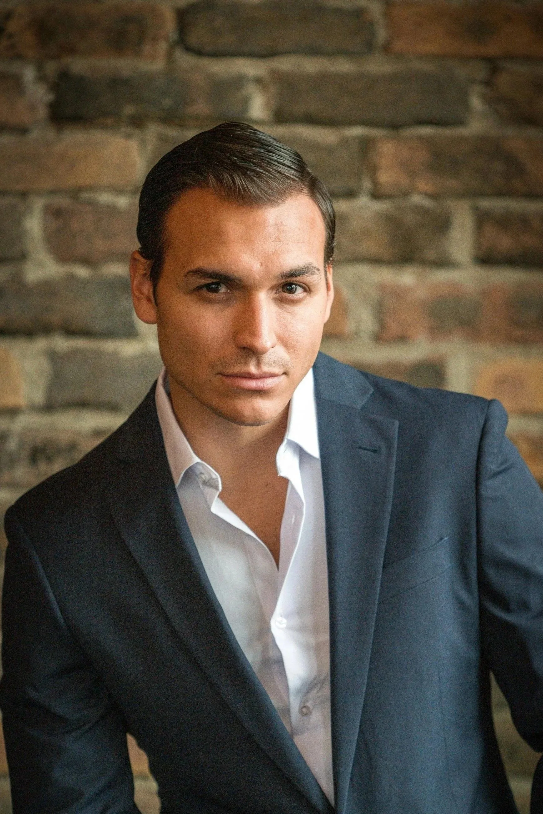 A man wearing a dark suit and white shirt, posing against a brick wall background.