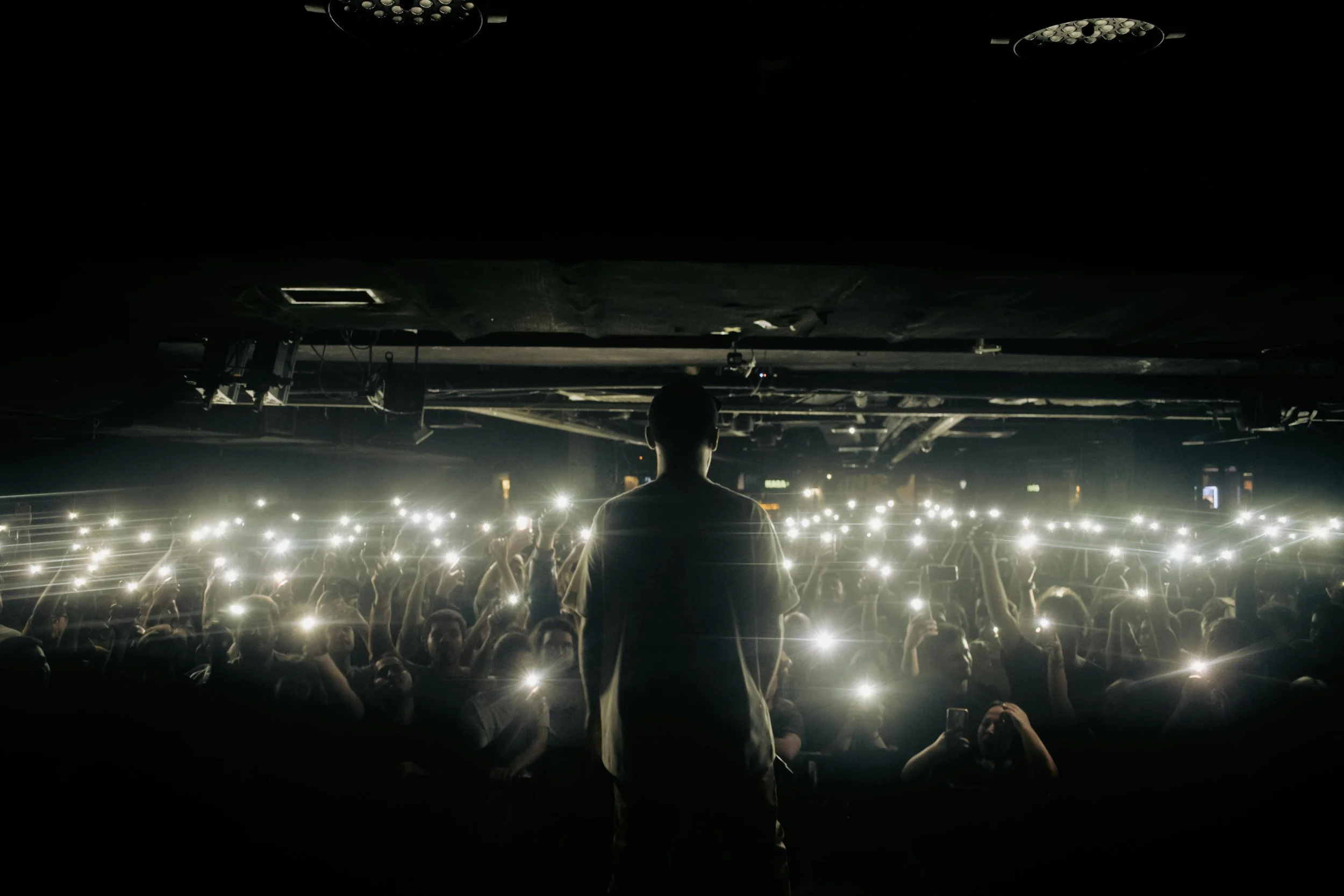 A person stands on stage silhouetted against a large crowd at a concert or event, with many audience members holding up their phones with flashlights on.