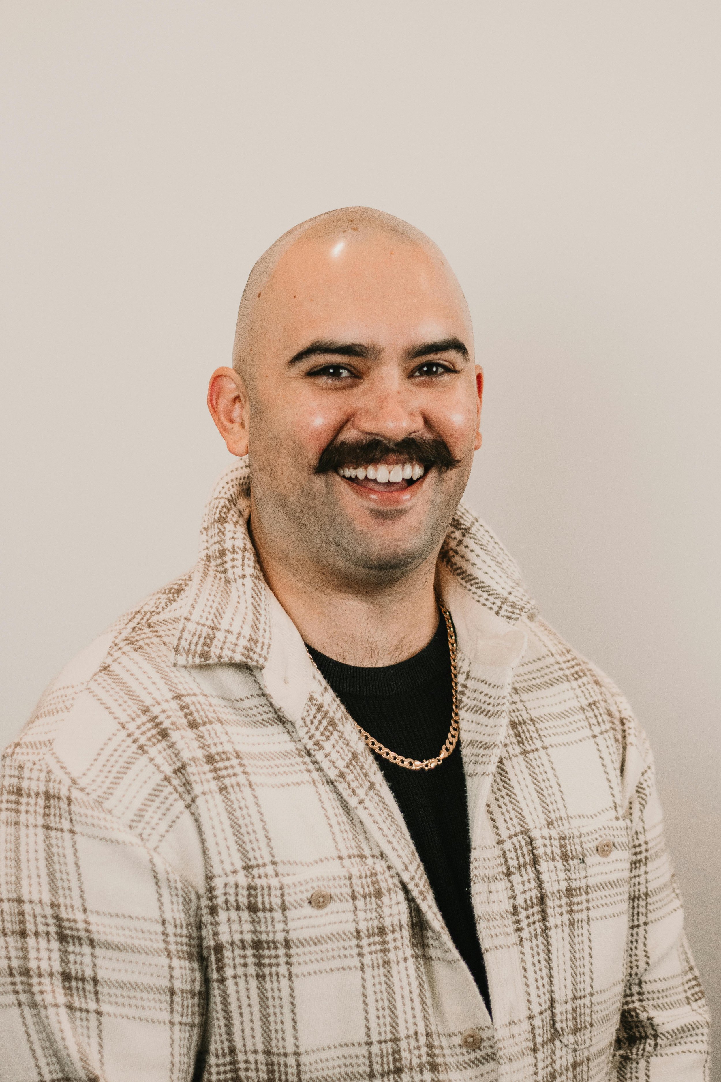 A smiling man with a shaved head, mustache, wearing a plaid jacket, black shirt, and gold chain, standing against a plain background.