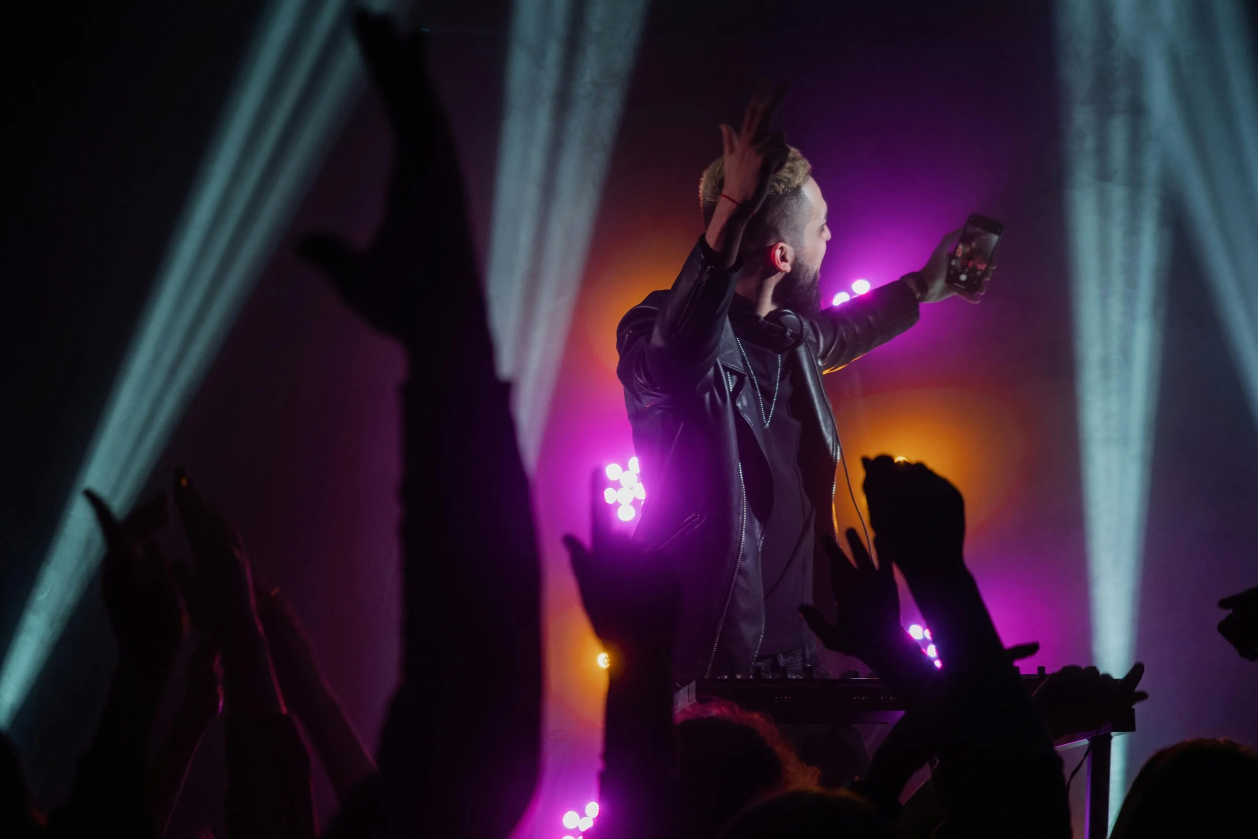 A DJ with dyed hair and a beard is performing at a concert, taking a selfie or recording a video with a smartphone while wearing headphones around his neck. There are colorful lights and an audience with raised hands in front of him.