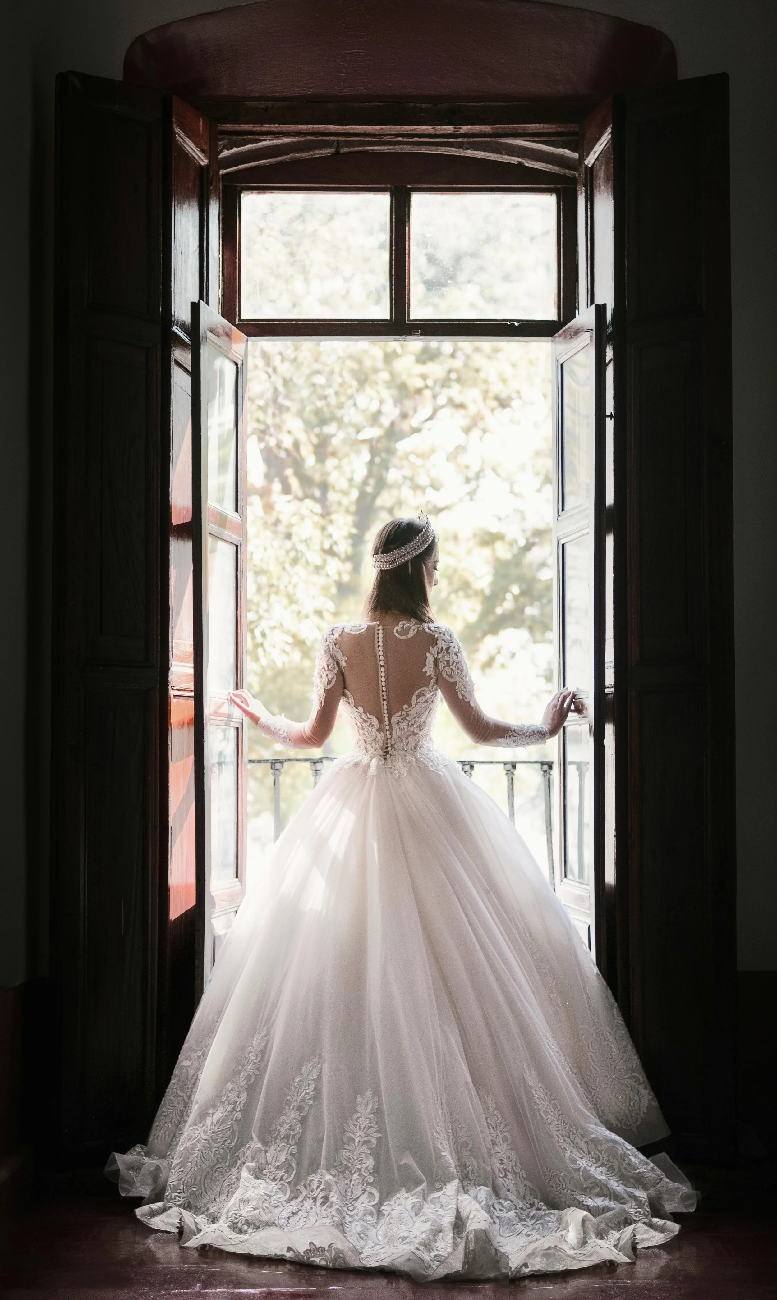 Bride in a white wedding gown standing at open window with back to camera, looking outside, with trees visible in the background.