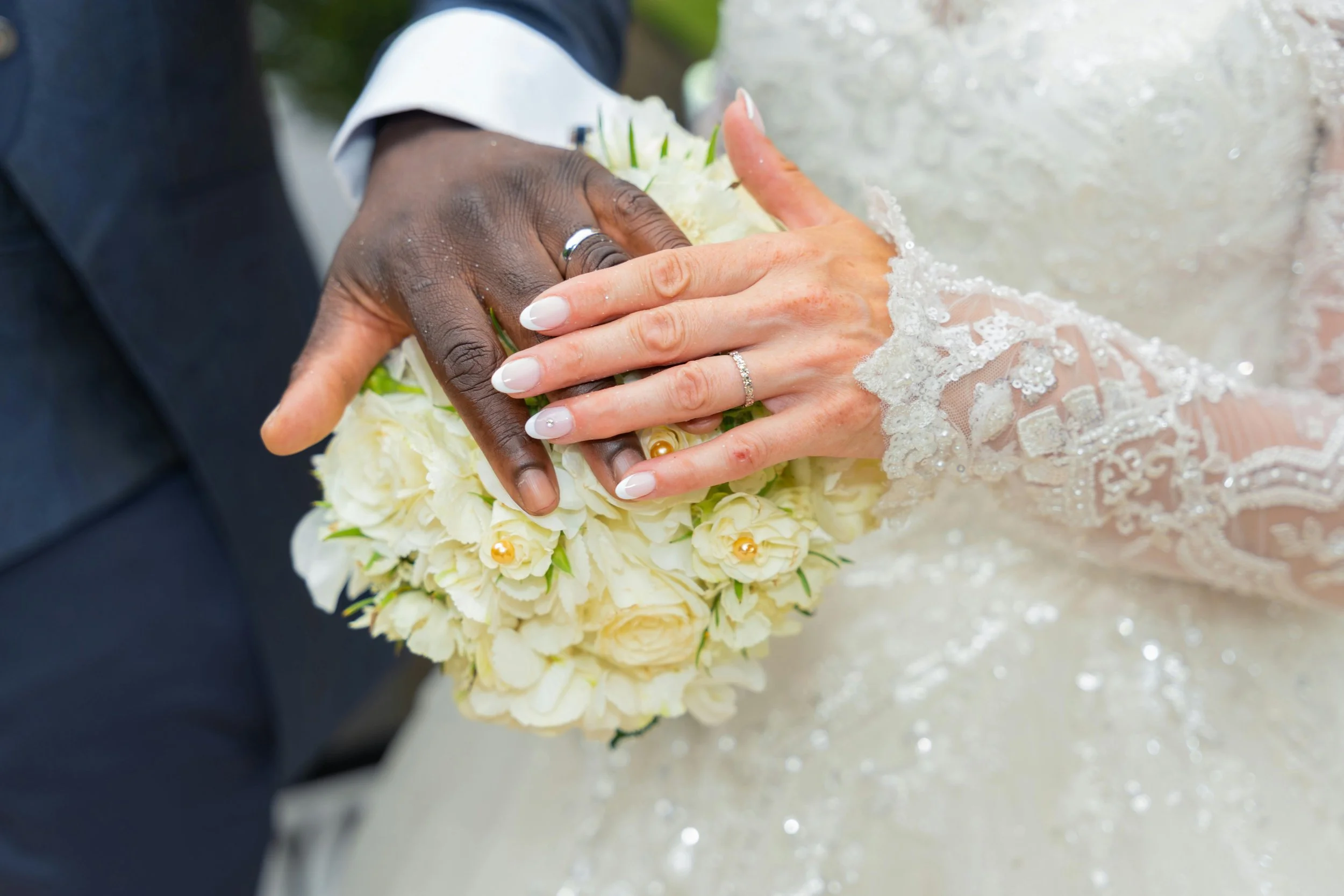 Close-up of a wedding couple's hands with wedding rings, holding a bouquet of white flowers, with the bride's lace sleeve visible.