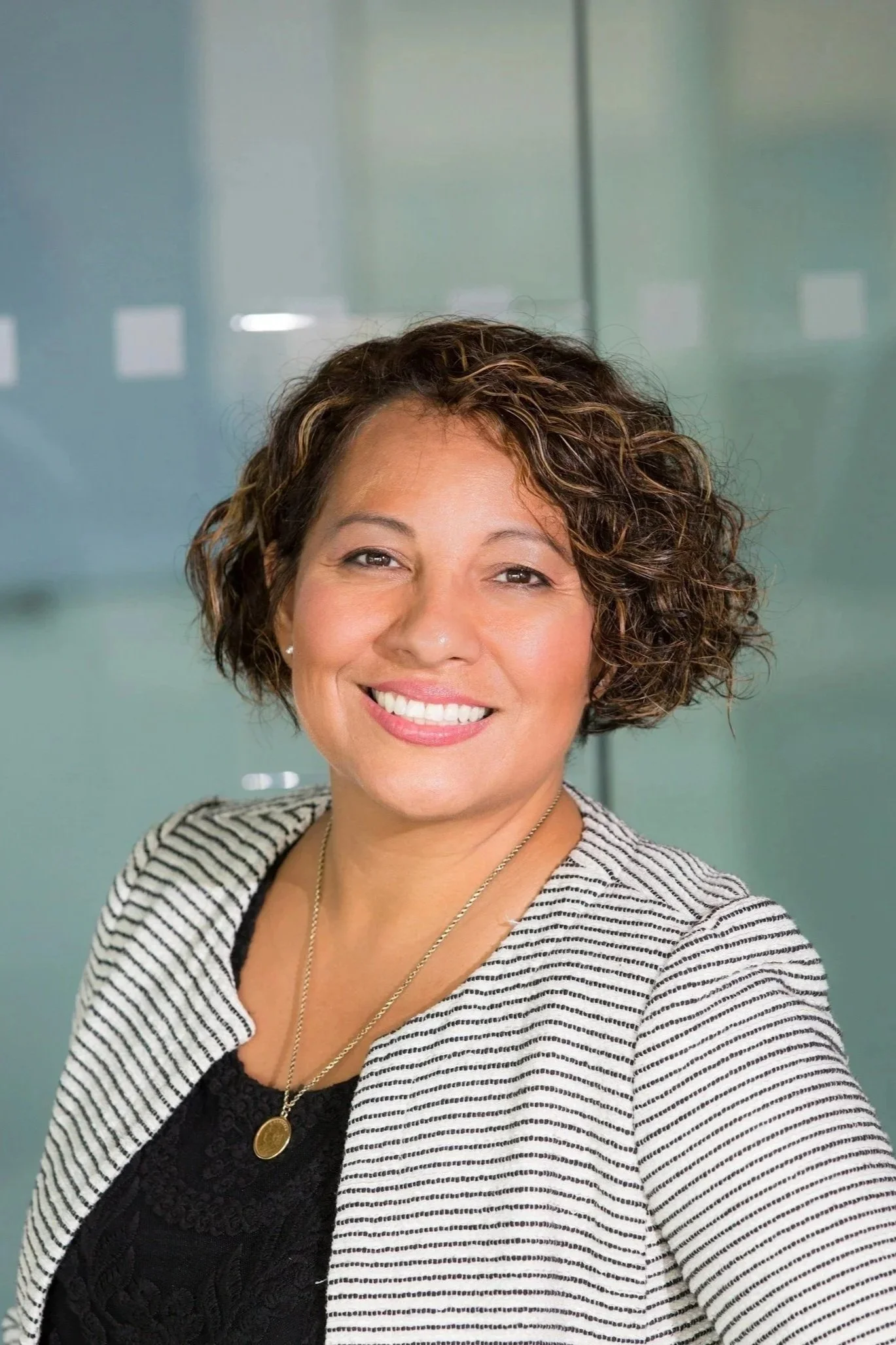 Smiling woman with curly brown hair, wearing a striped blazer, black top, and a gold necklace, standing in front of a glass wall.
