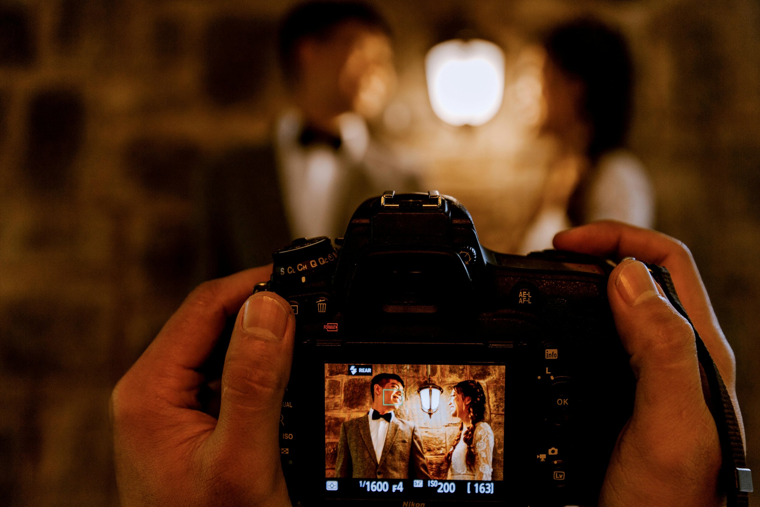 Photographer taking a picture of a couple dressed in vintage formal attire, with the camera's screen showing their smiling faces.
