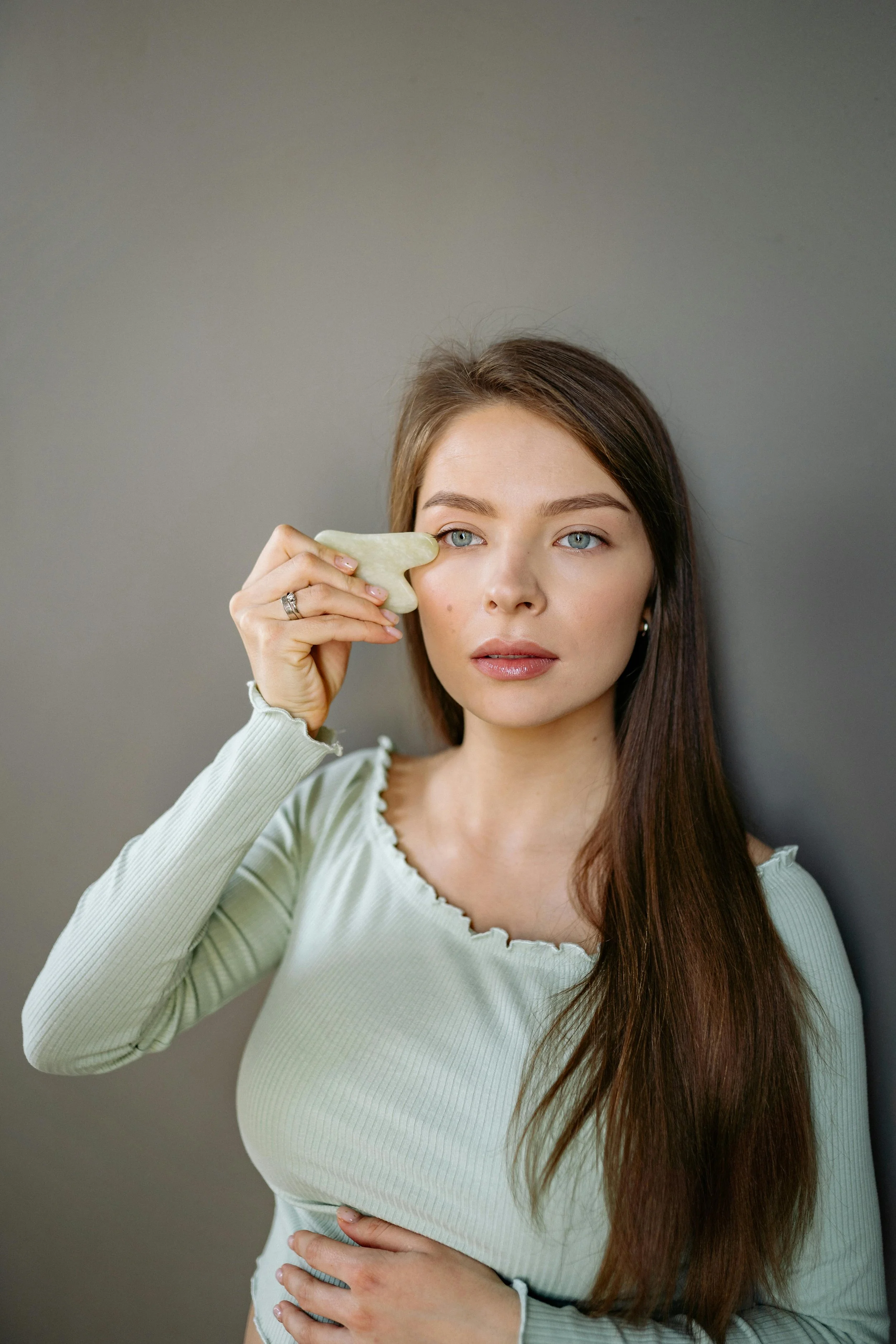 A woman with long brown hair and fair skin holds a jade roller to her cheek while standing against a gray wall.