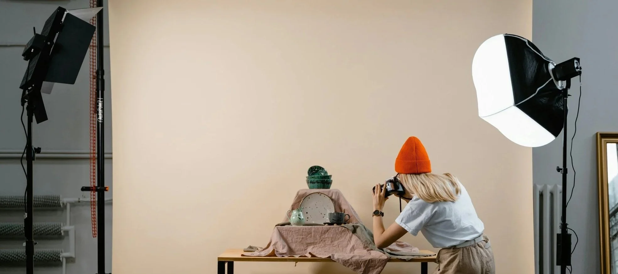 A woman with blonde hair, wearing an orange beanie, white t-shirt, and beige pants, is taking a photograph of a still life arrangement in a photography studio. The setup includes a ceramic pitcher, bowls, and cups on a table covered with cloth. The studio has professional lighting equipment, with a large softbox on the right and a smaller light on the left.