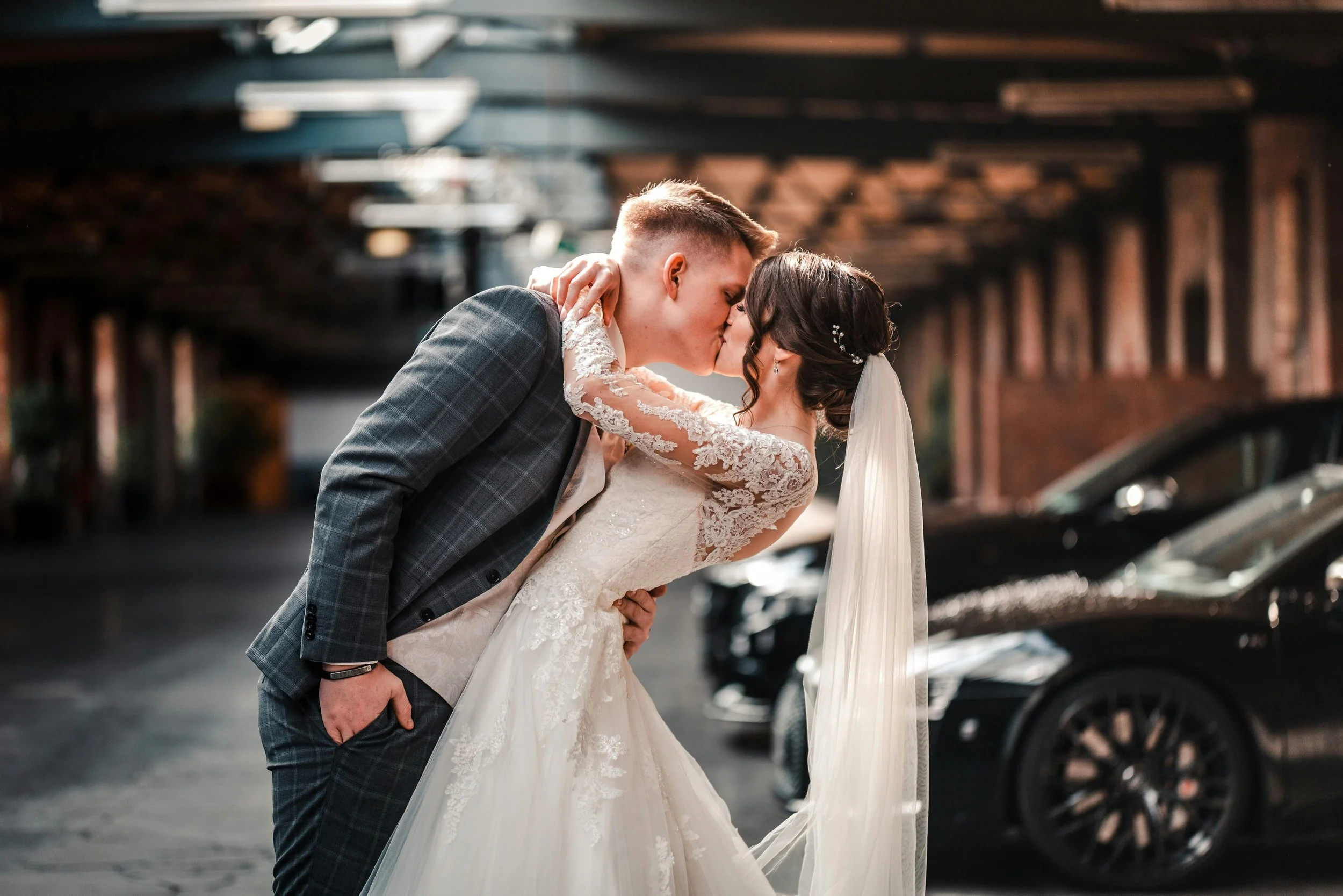 A bride and groom sharing a kiss in a parking garage, with the groom leaning forward and the bride holding his neck, both dressed in wedding attire.