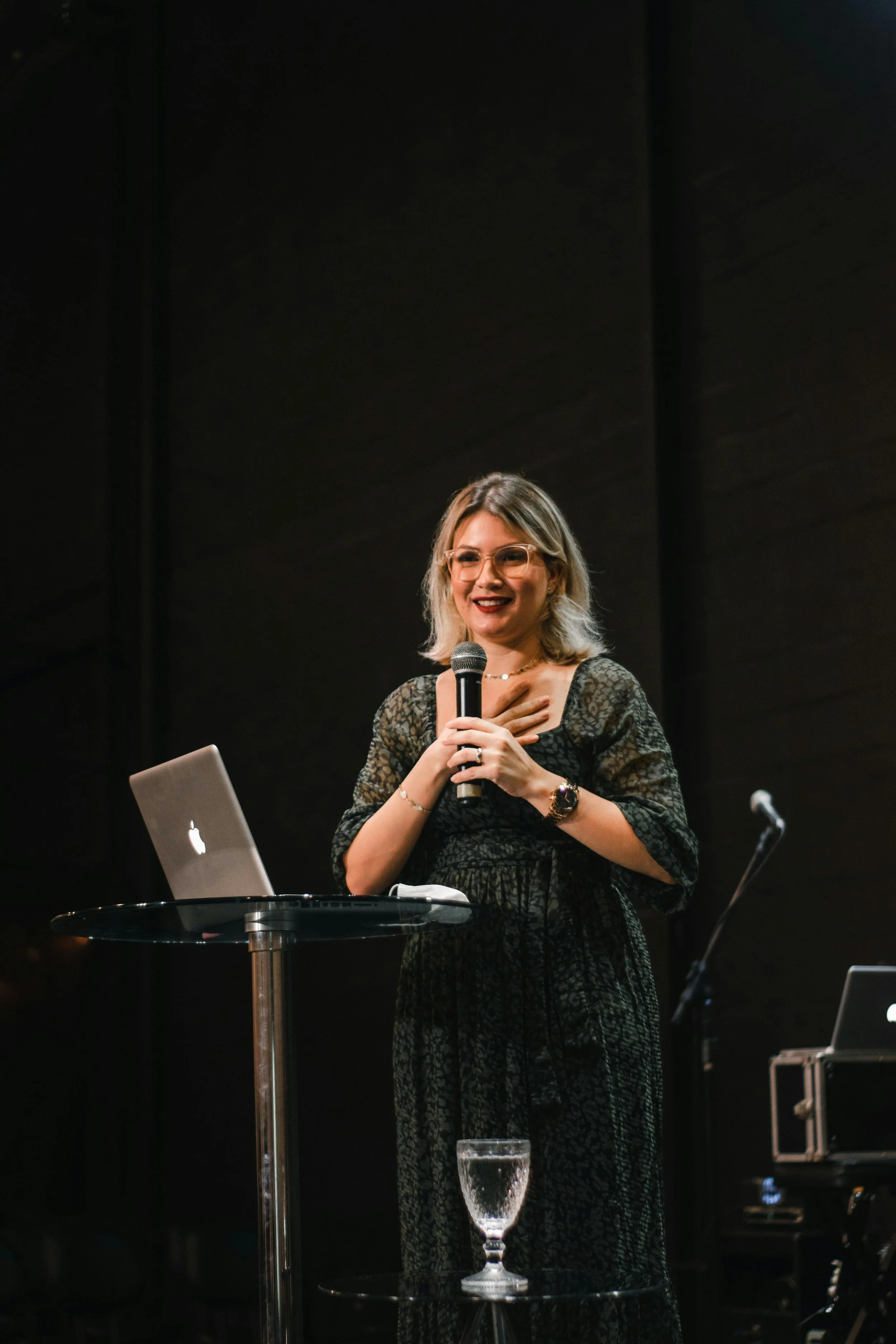 A woman with blonde hair and wearing glasses, holding a microphone and speaking at a podium with a laptop, in a dark room with a black background.