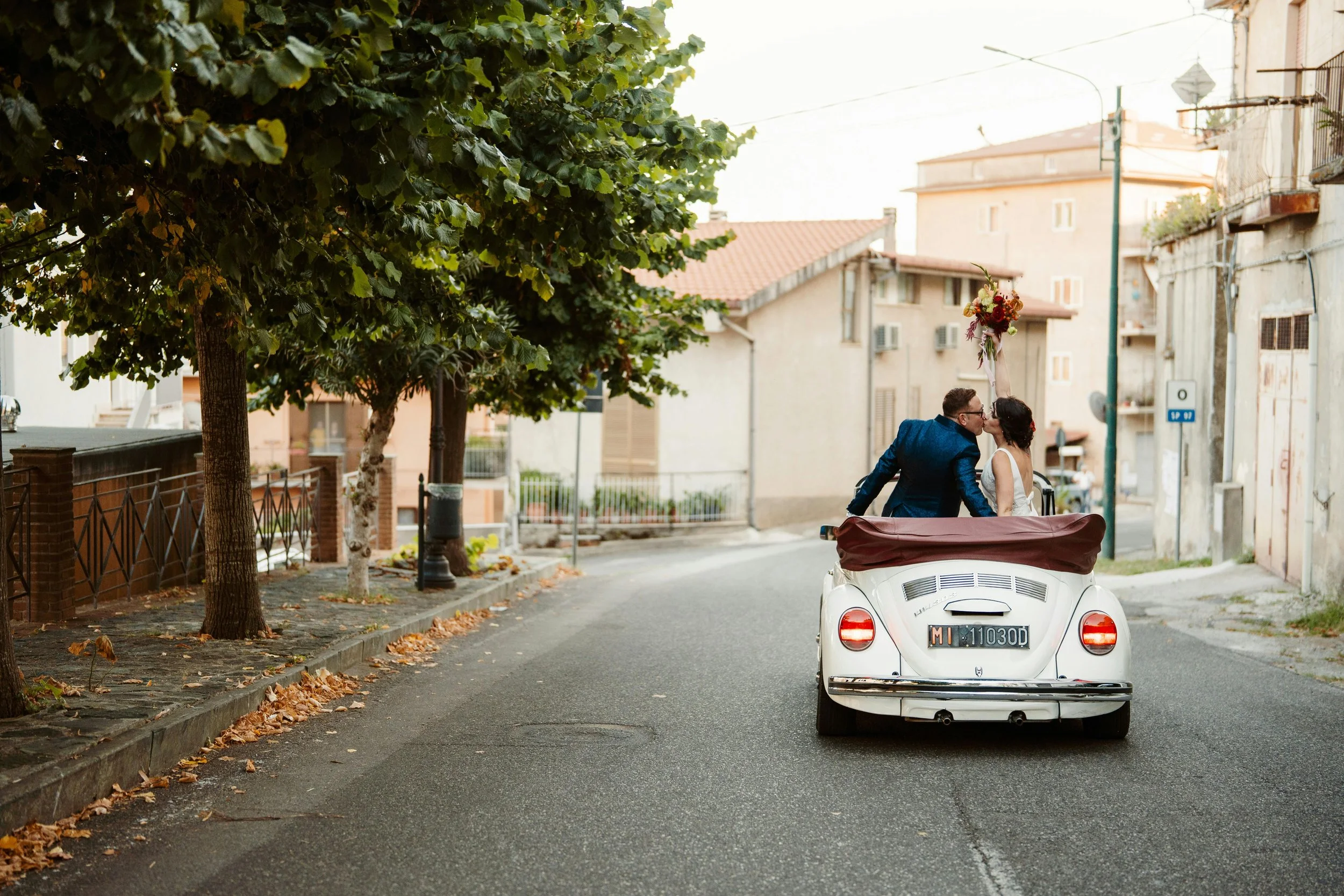A newlywed couple sharing a kiss while riding in a classic white convertible car on a quiet city street with trees and buildings.