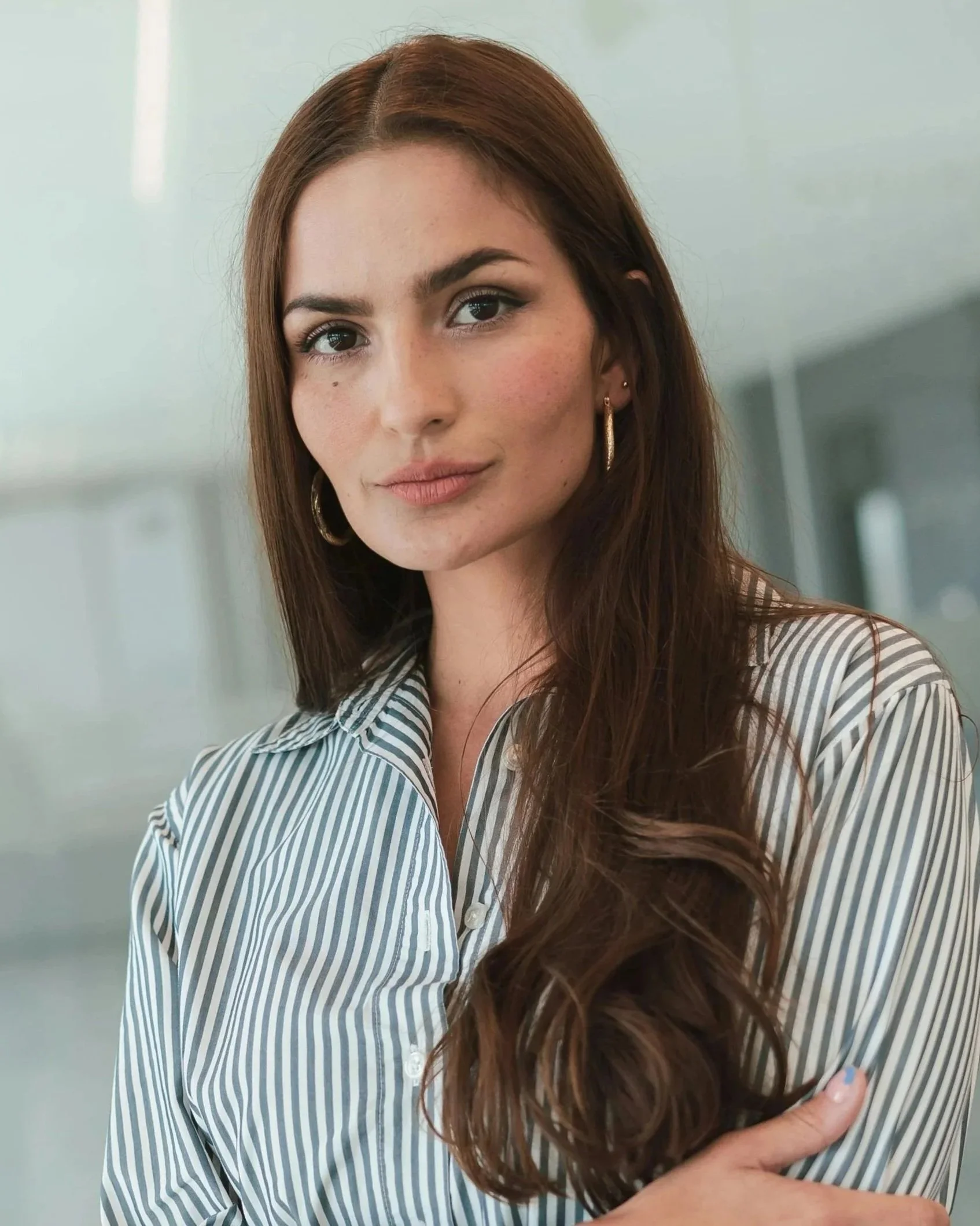 A young woman with long brown hair, wearing a striped button-up shirt and gold hoop earrings, looking at the camera indoors.