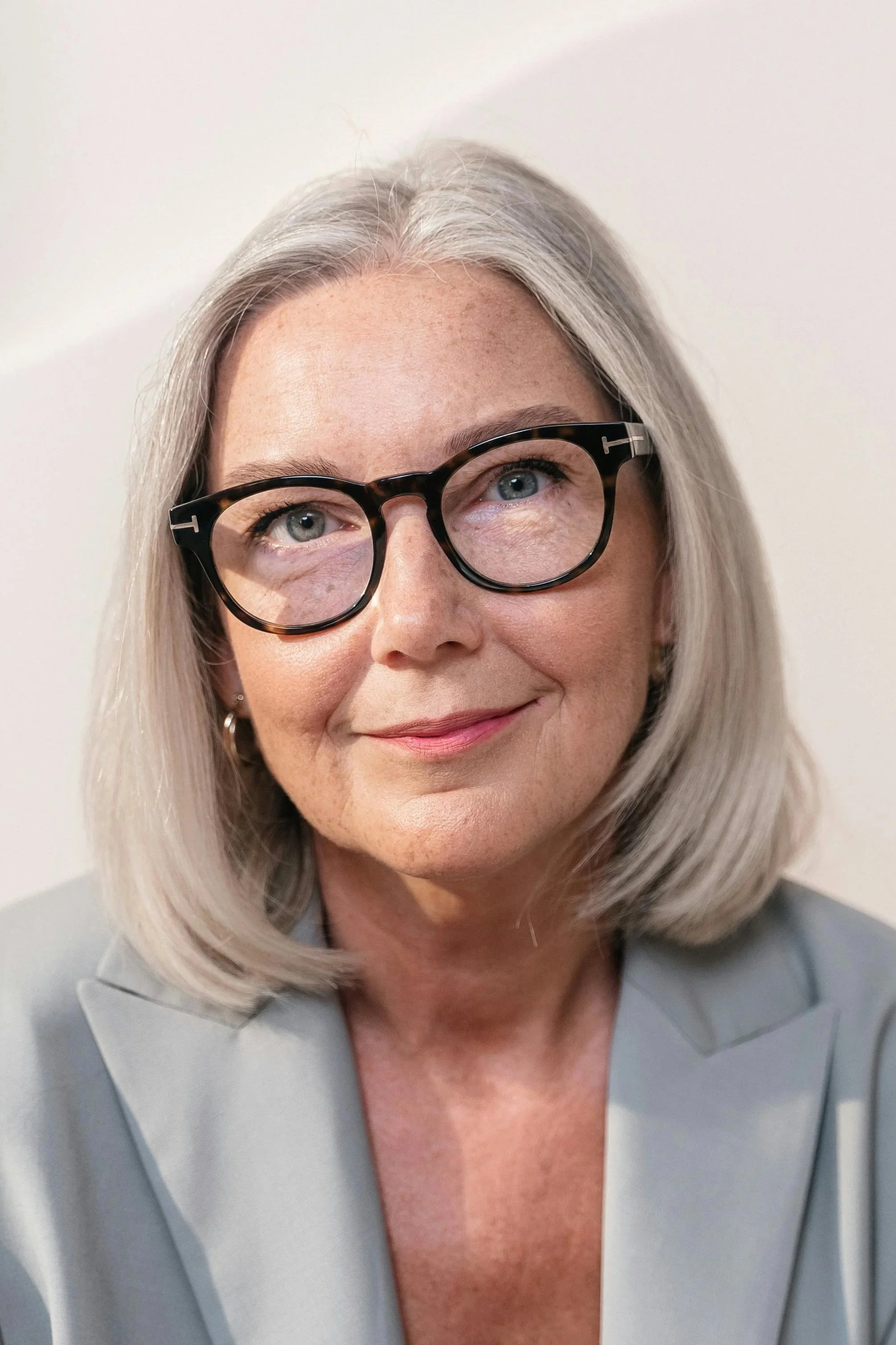 A smiling woman with shoulder-length gray hair, wearing black glasses and a light gray blazer, against a plain light background.
