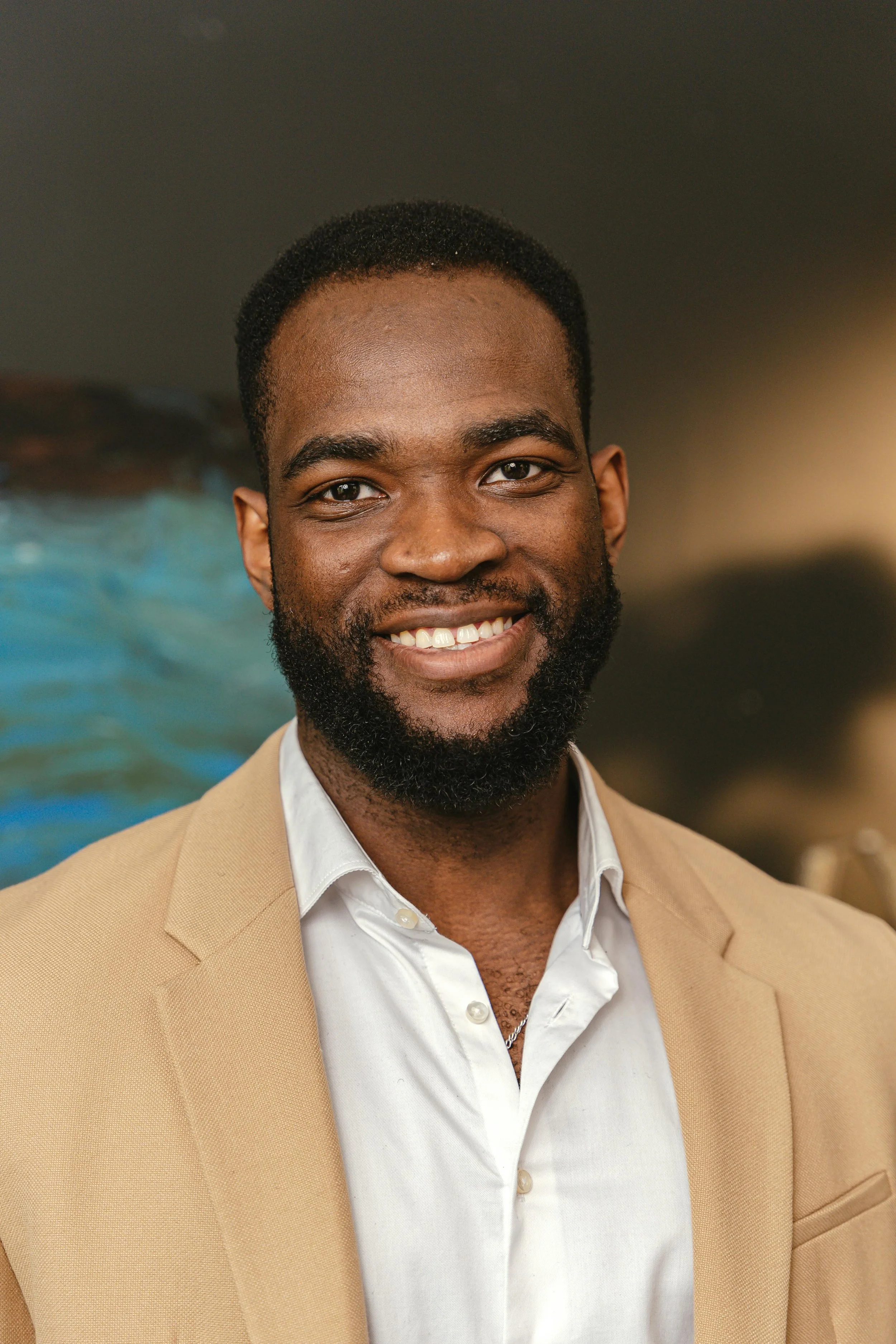 Portrait of a smiling man with a beard wearing a beige blazer over a white shirt, standing indoors with a blurred background.