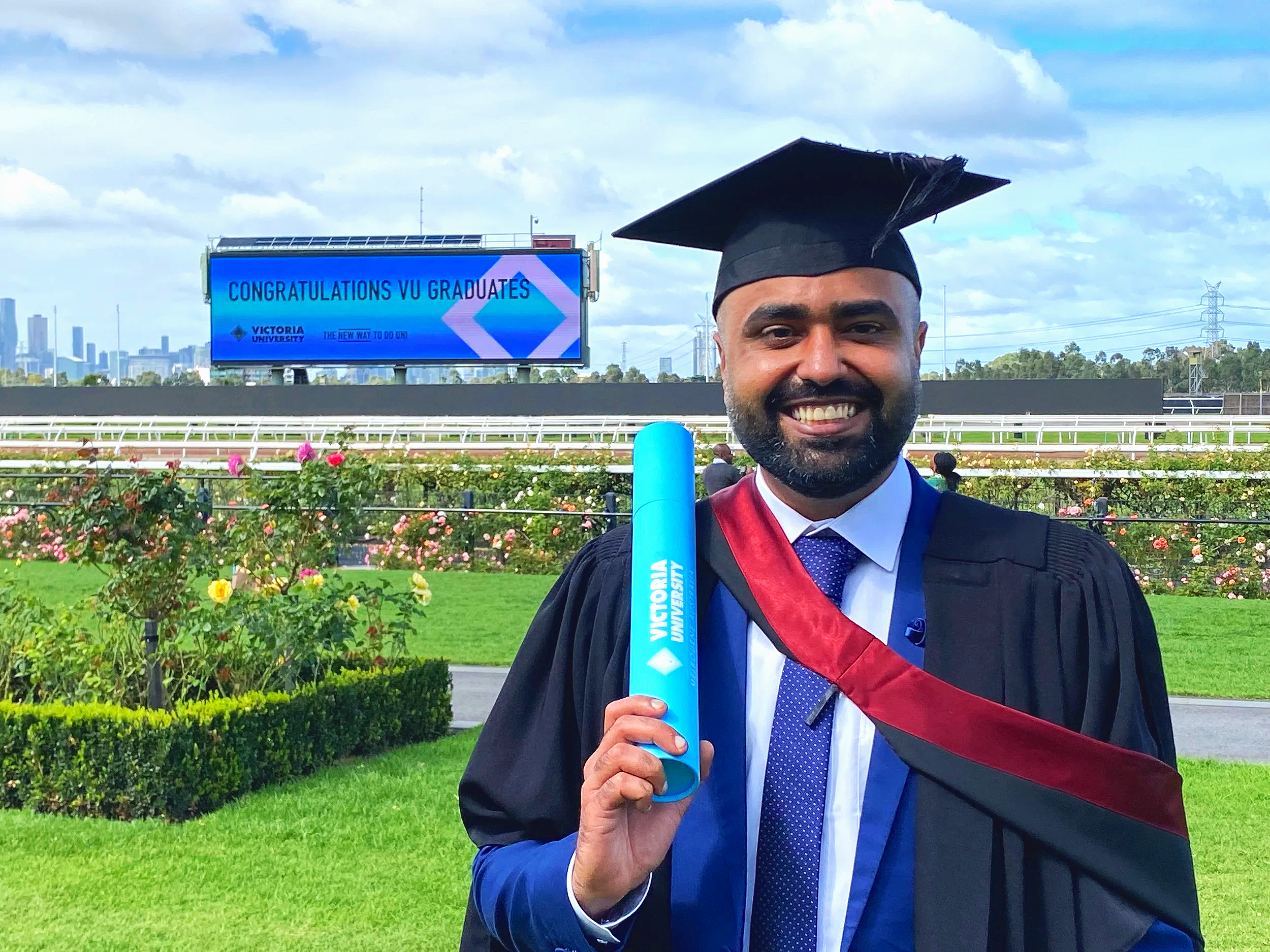 A man in graduation attire, including a cap and gown, holding a diploma in front of a backdrop of rose bushes and a digital screen that reads 'Congratulations VU Graduates', with a city skyline in the distance.