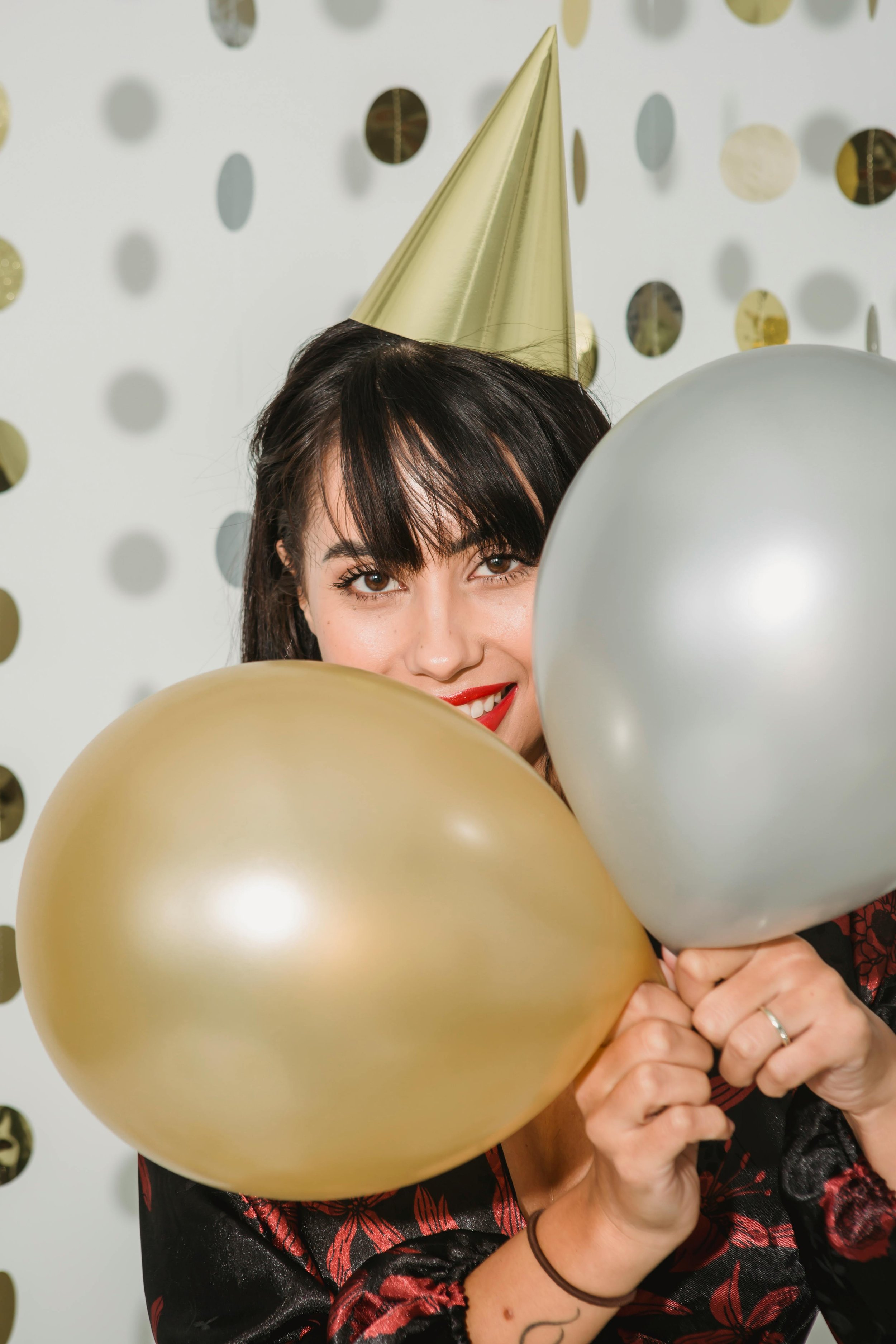 A woman wearing a gold party hat and red lipstick, holding gold and silver balloons, at a celebration with gold and silver decor in the background.
