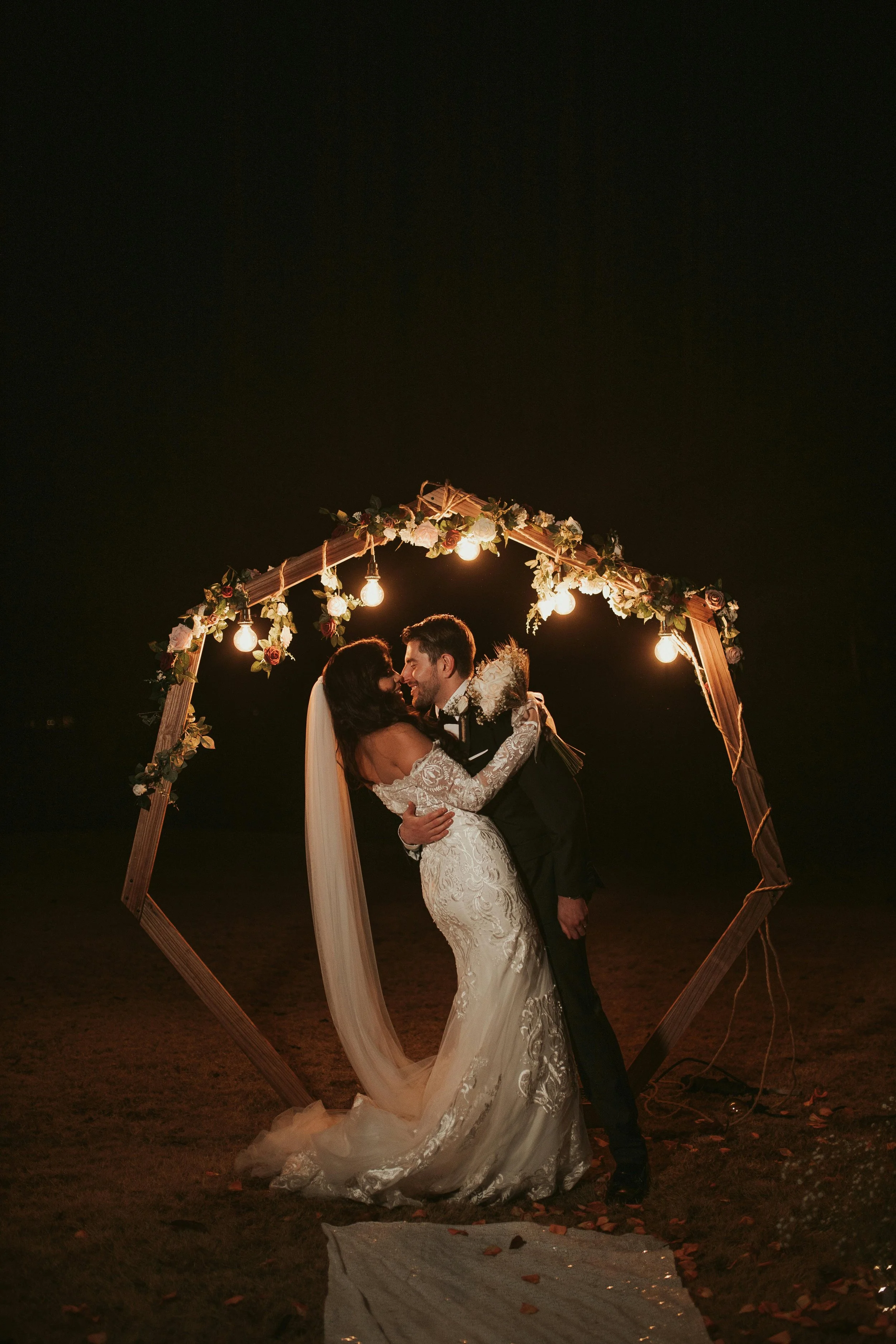 A bride and groom share a kiss at night under a decorated wooden arch with hanging lights during their outdoor wedding celebration.