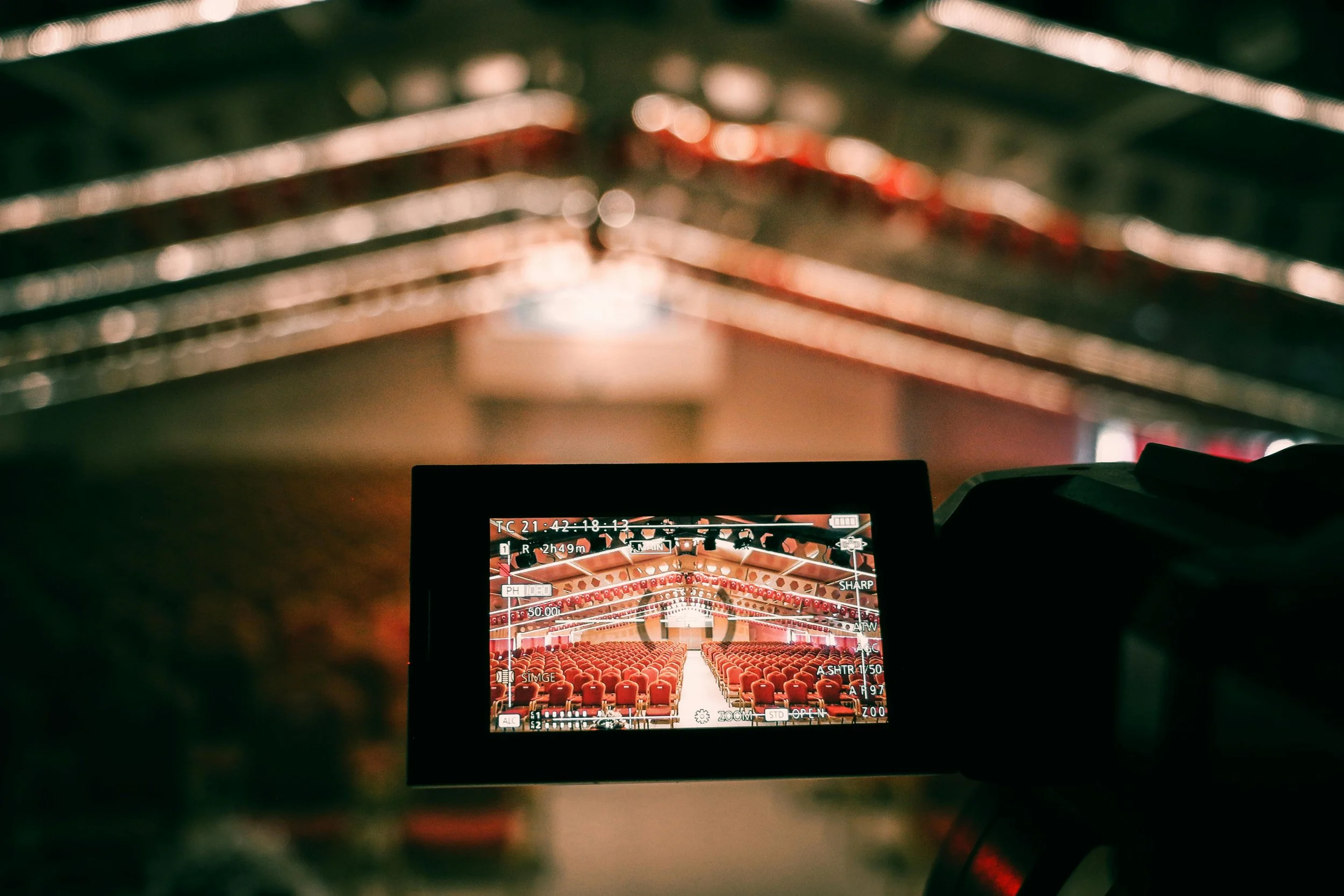 Interior view of a theater or auditorium with rows of red seats, photographed through a camera monitor in a dimly lit setting with a blurred background.