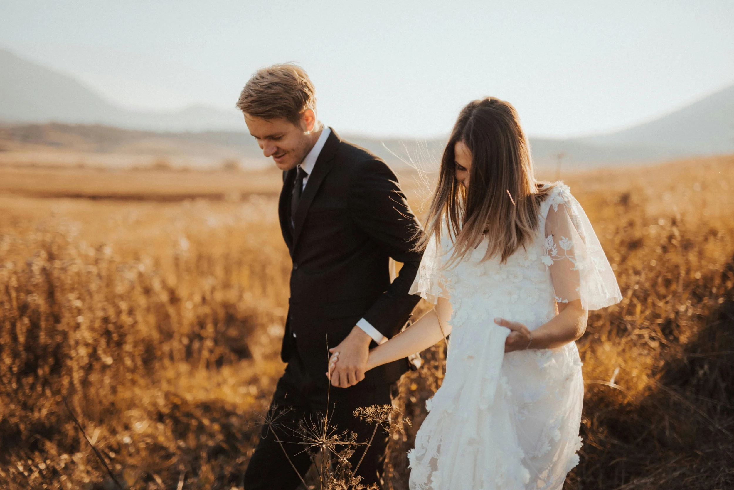 A couple dressed in wedding attire walking hand-in-hand through a golden field at sunset.