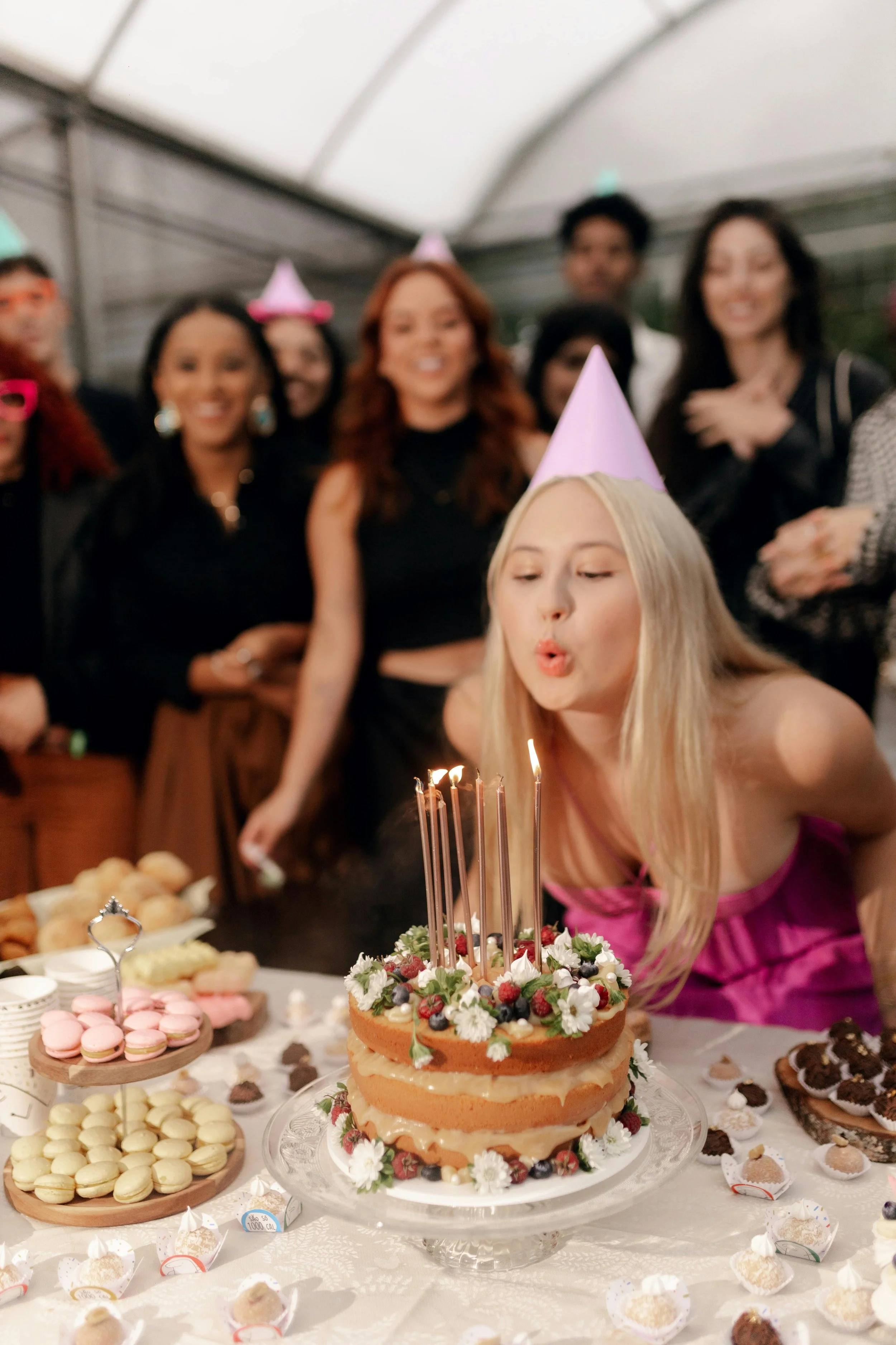 A woman in a pink dress blows out candles on a birthday cake decorated with flowers and berries, surrounded by friends at a party.