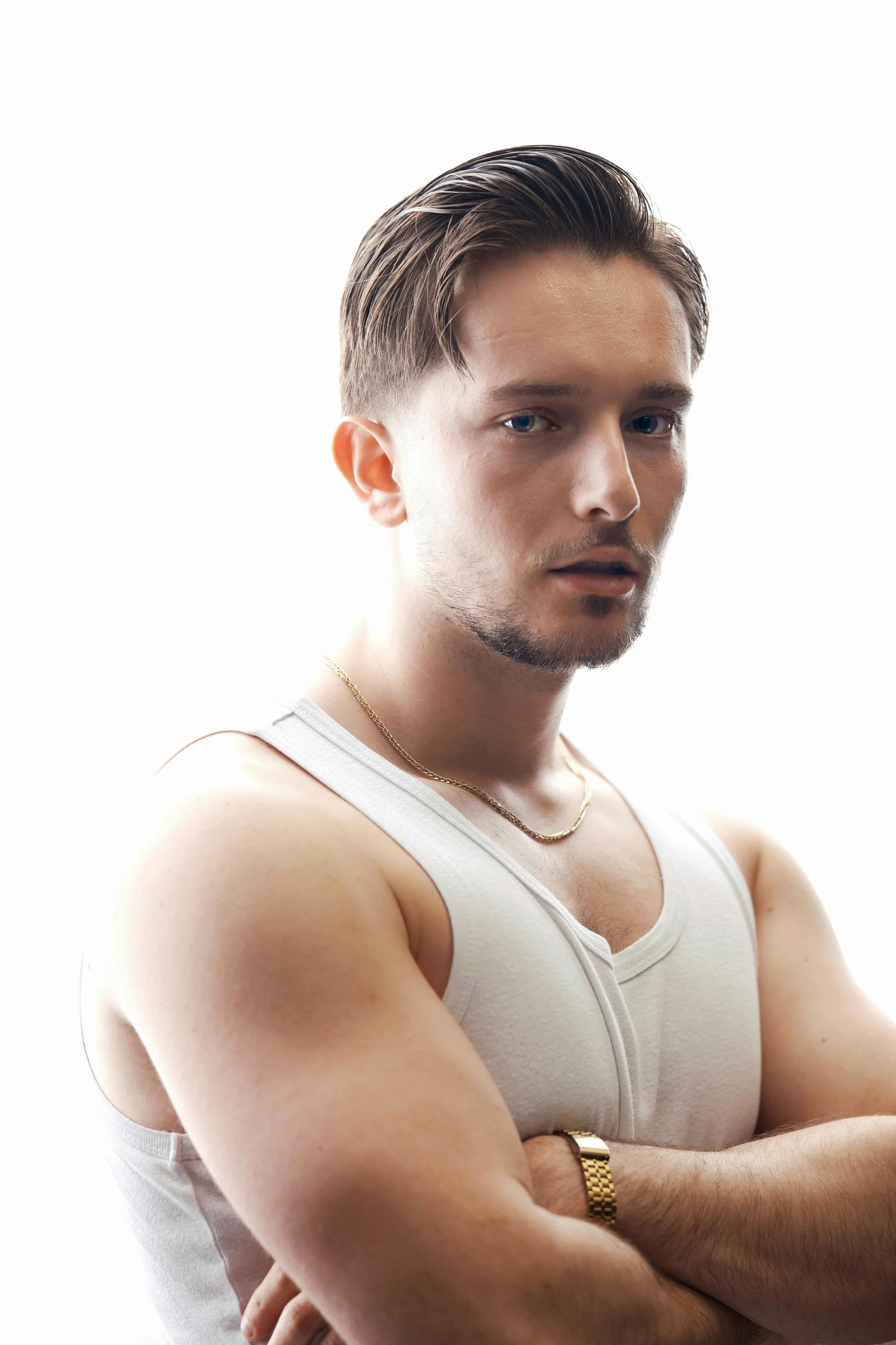 A young man with styled hair and a light beard, wearing a white tank top, gold chain, and watch, poses with arms crossed against a plain white background.