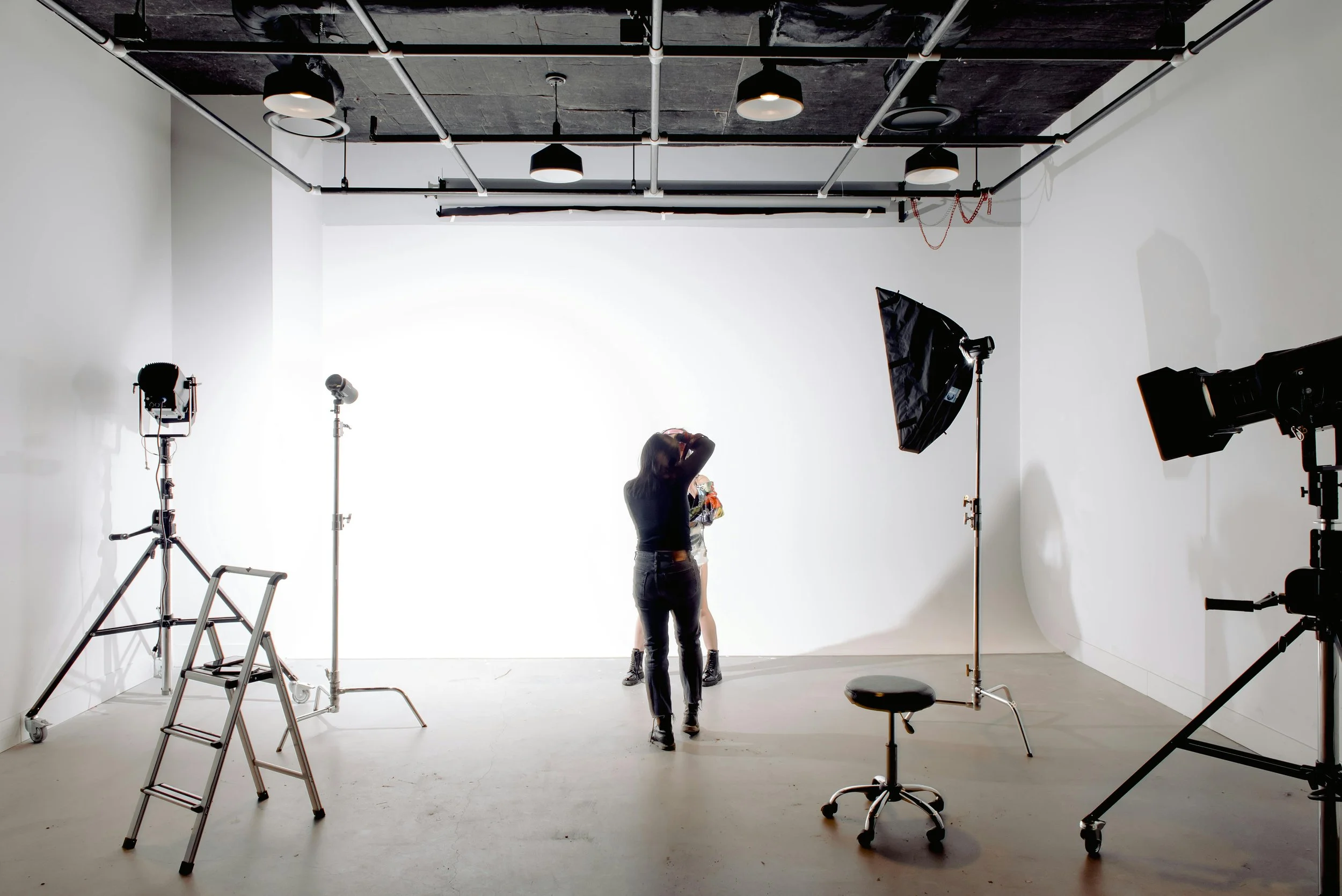 A photography studio with a white backdrop, professional lighting equipment, and two people in the center preparing for a photo shoot.