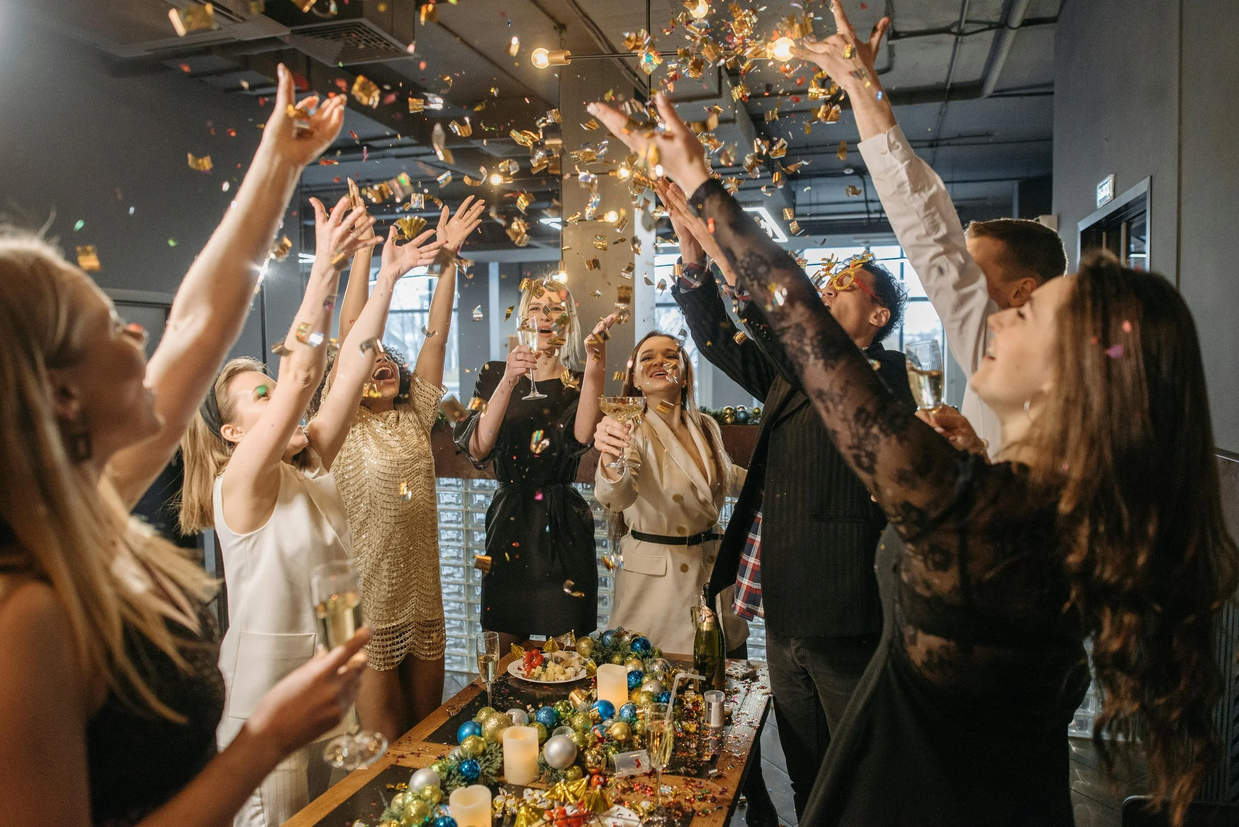 People celebrating at a festive party, throwing confetti and holding drinks around a decorated table with ornaments and candles.