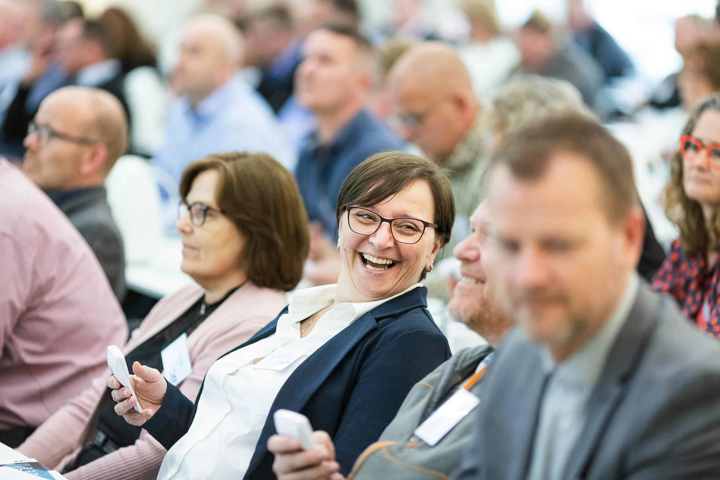People attending a conference, some using their phones, with a woman laughing and sitting in the center.