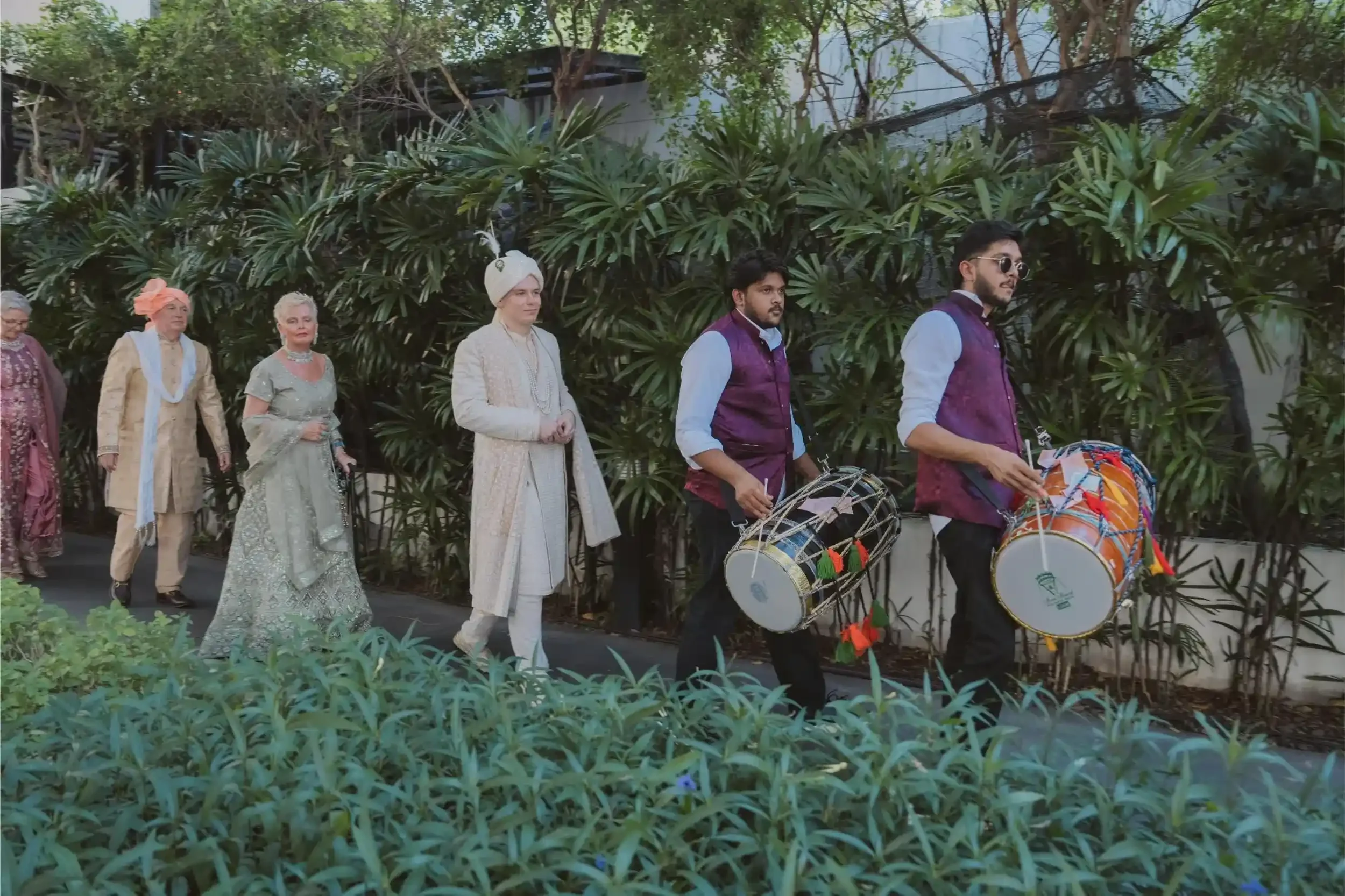 A group of people dressed in traditional and modern clothing participating in a cultural parade with drummers, walking along a path surrounded by lush green plants and trees.