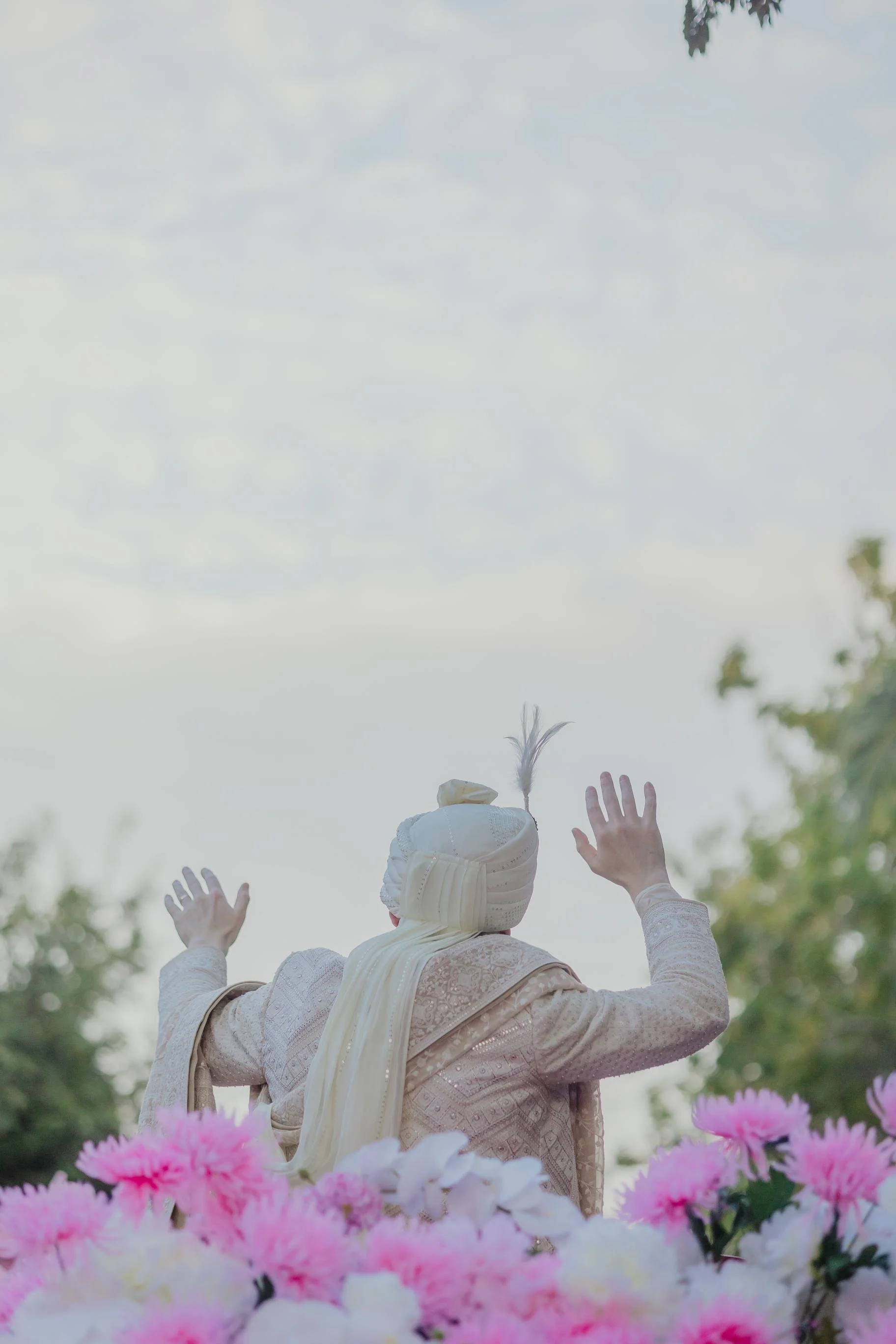 Groom dressed in elegant clothing and head covering raising hands, surrounded by pink and white flowers, with trees and cloudy sky in the background.