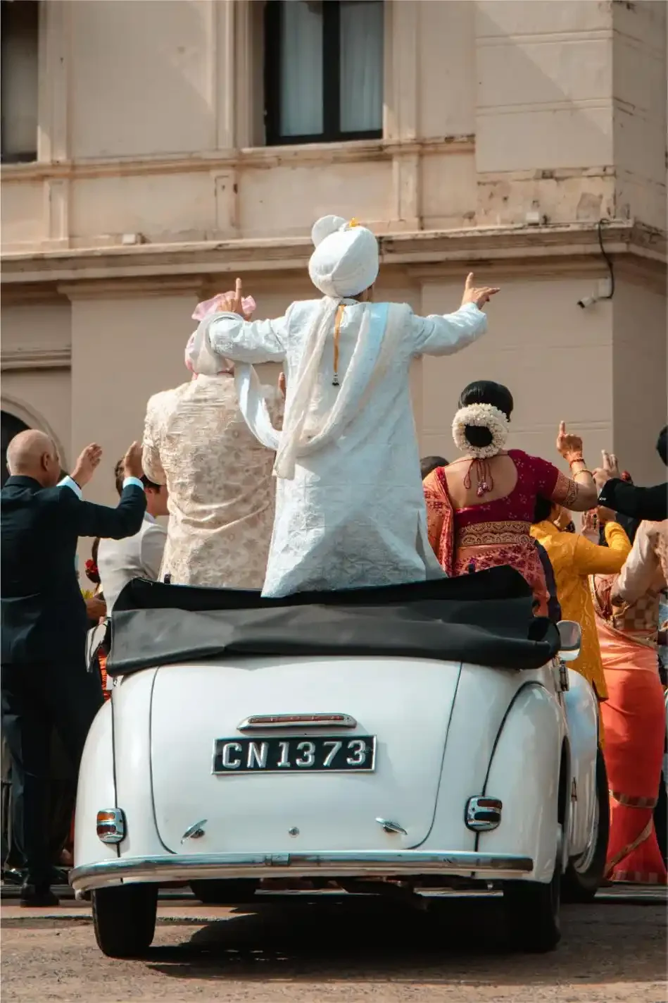 People celebrating on a white vintage car during a cultural parade, with traditional attire and festive atmosphere.