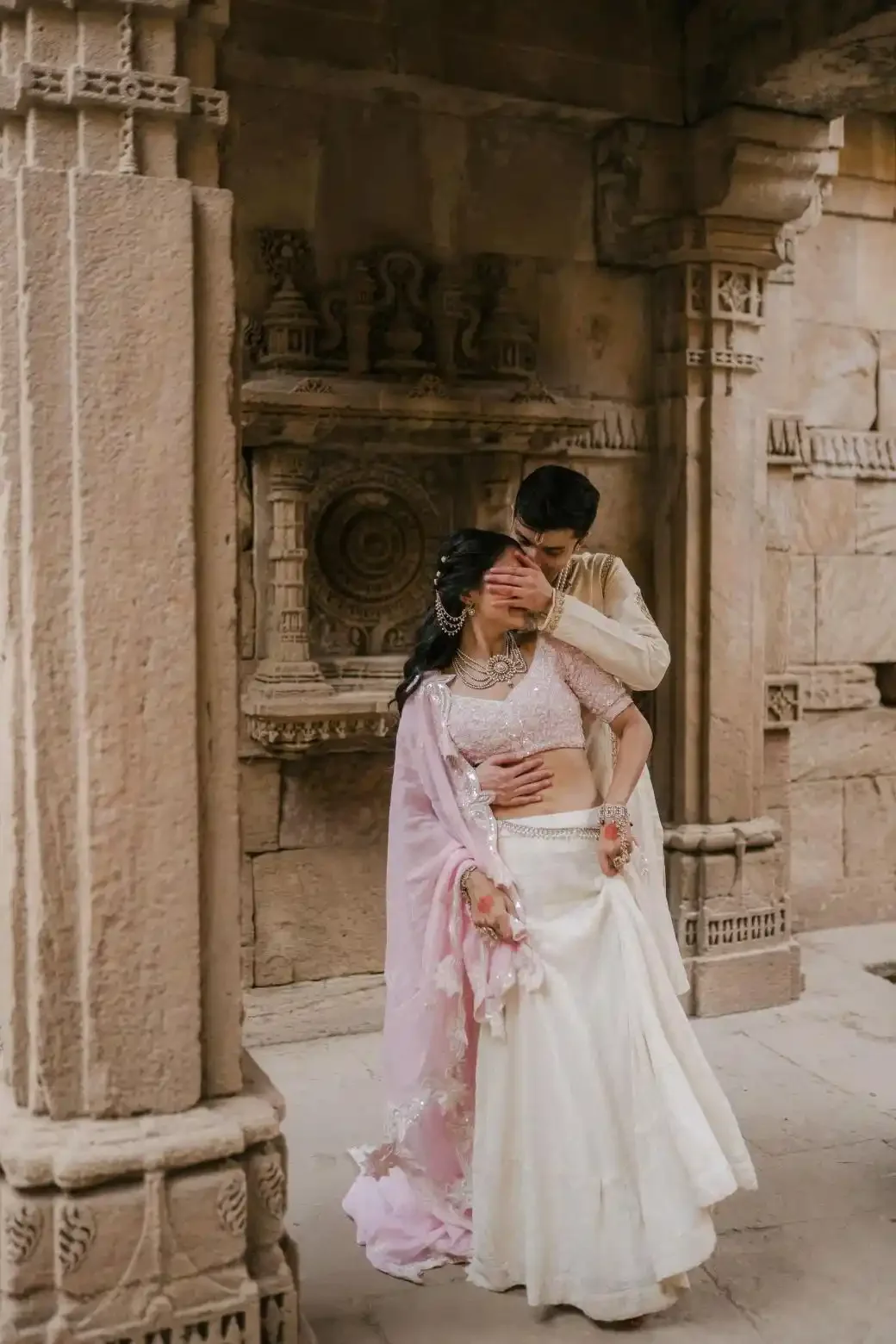 A couple dressed in traditional Indian wedding attire sharing an intimate moment inside an ancient stone temple with intricate carvings.