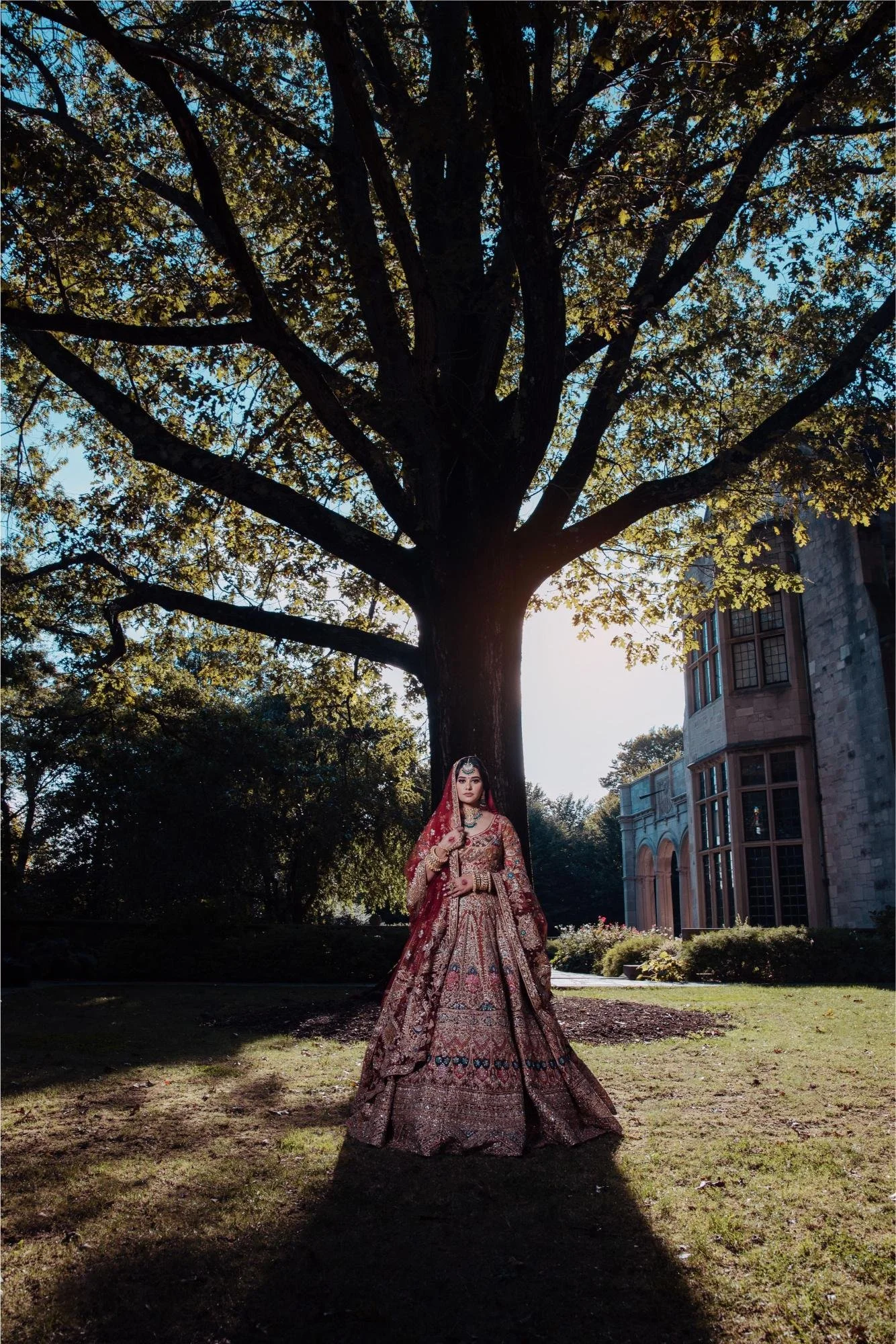 A woman in ornate traditional Indian bridal attire, a heavily embroidered maroon and gold lehenga, standing outdoors under a large tree with sprawling branches and leaves, with a historic stone building in the background.