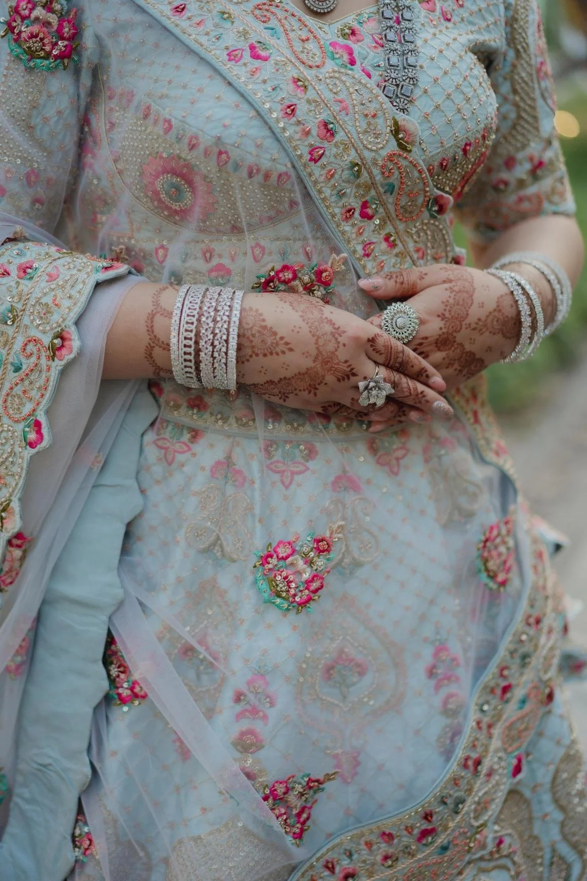 Close-up of a bride wearing a traditional, heavily embroidered white and pink wedding dress with intricate floral patterns, jewelry, and henna on her hands.