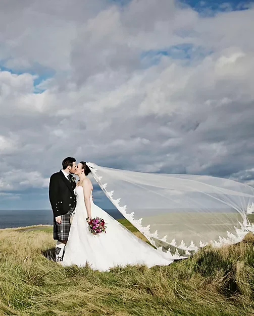 A bride and groom standing close together on a grassy hilltop, about to kiss, with a cloudy sky and ocean in the background. The bride is holding a bouquet, and her veil is flowing in the wind.