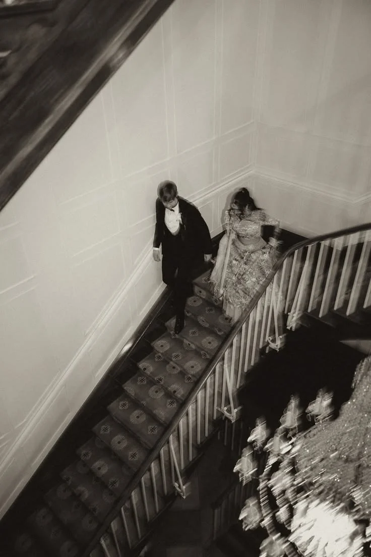 A black and white photo of a man and woman dressed in formal attire walking up a spiral staircase in an elegant setting, viewed from a top angle.