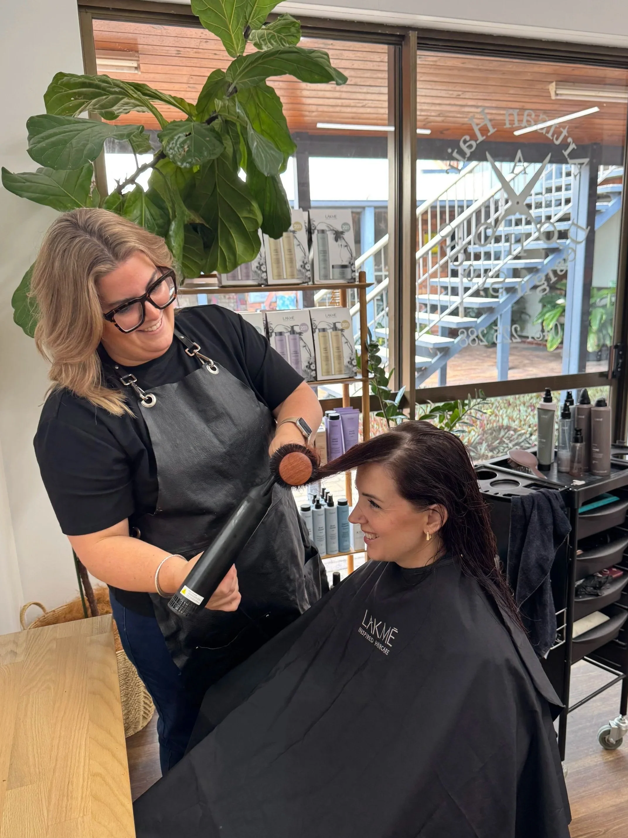 A hairstylist is blow-drying a woman's dark hair in a salon. The woman is sitting in a chair with a black cape that says 'LAKME INSPIRED HAIRCARE.' The stylist is smiling, holding a round hairbrush and a blow dryer. The salon has large windows, a tall green plant, and shelves with hair products.