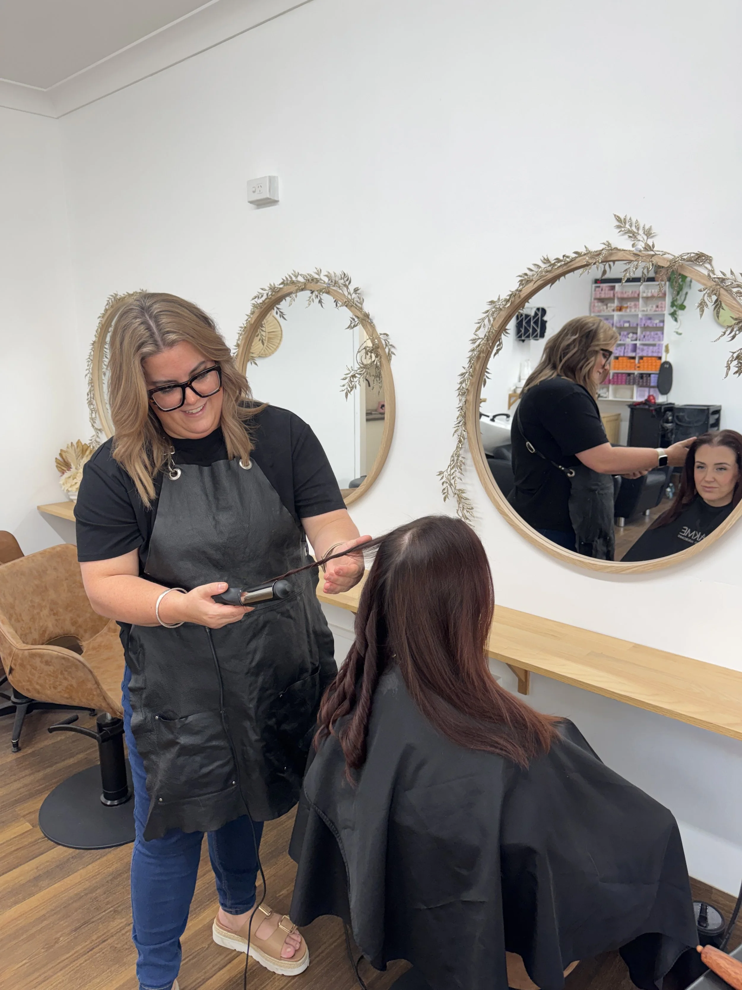 A woman with blonde hair and glasses is styling another woman's hair with a curling iron in a salon. The client has long, auburn hair and is seated in a styling chair. The salon wall has three round mirrors with decorative frames, and a small section of the salon's interior is visible.