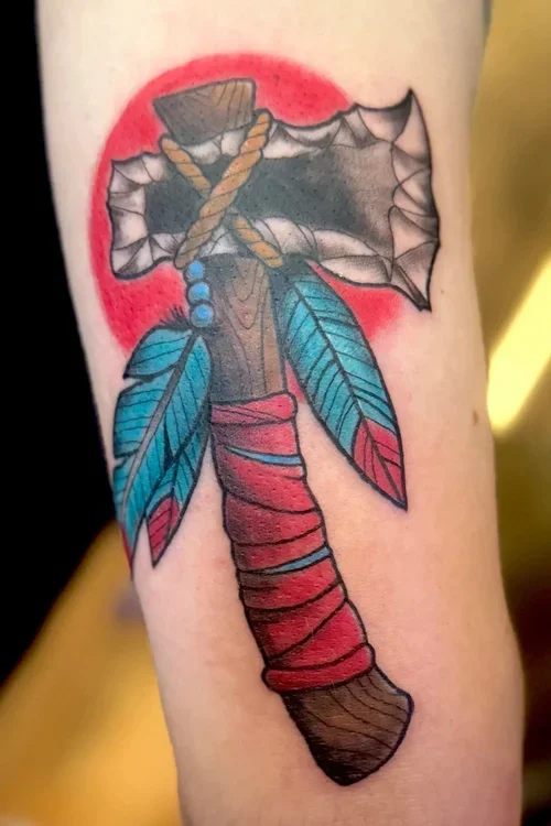 Tattoo of a traditional style axe with blue and red feathers and a red spot behind it.