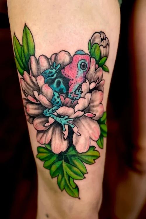 A colorful tattoo of a pink and blue frog resting on pink peony flowers with green leaves, covering the upper thigh.