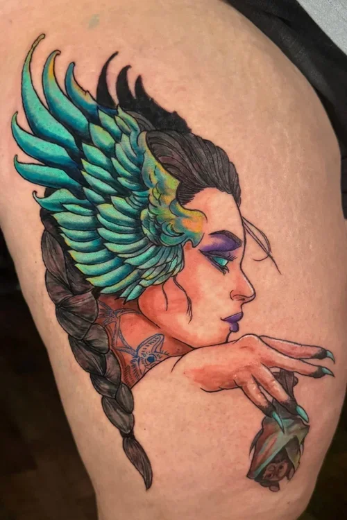 A colorful tattoo of a woman with closed eyes and a long braid, featuring a headdress of vibrant feathers and her hand holding a rose.