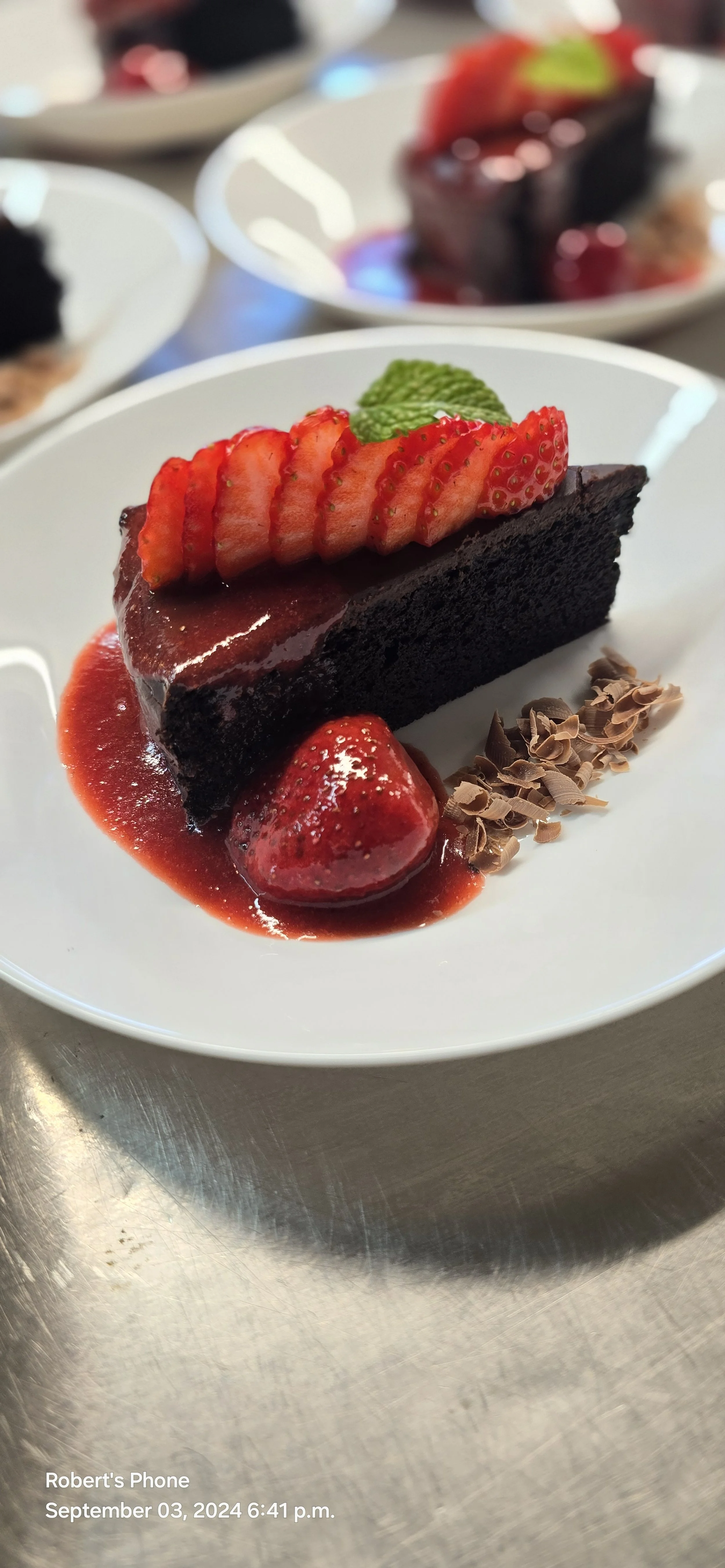 A slice of chocolate cake with strawberries and mint on top, served with strawberry sauce and chocolate shavings on a white plate.