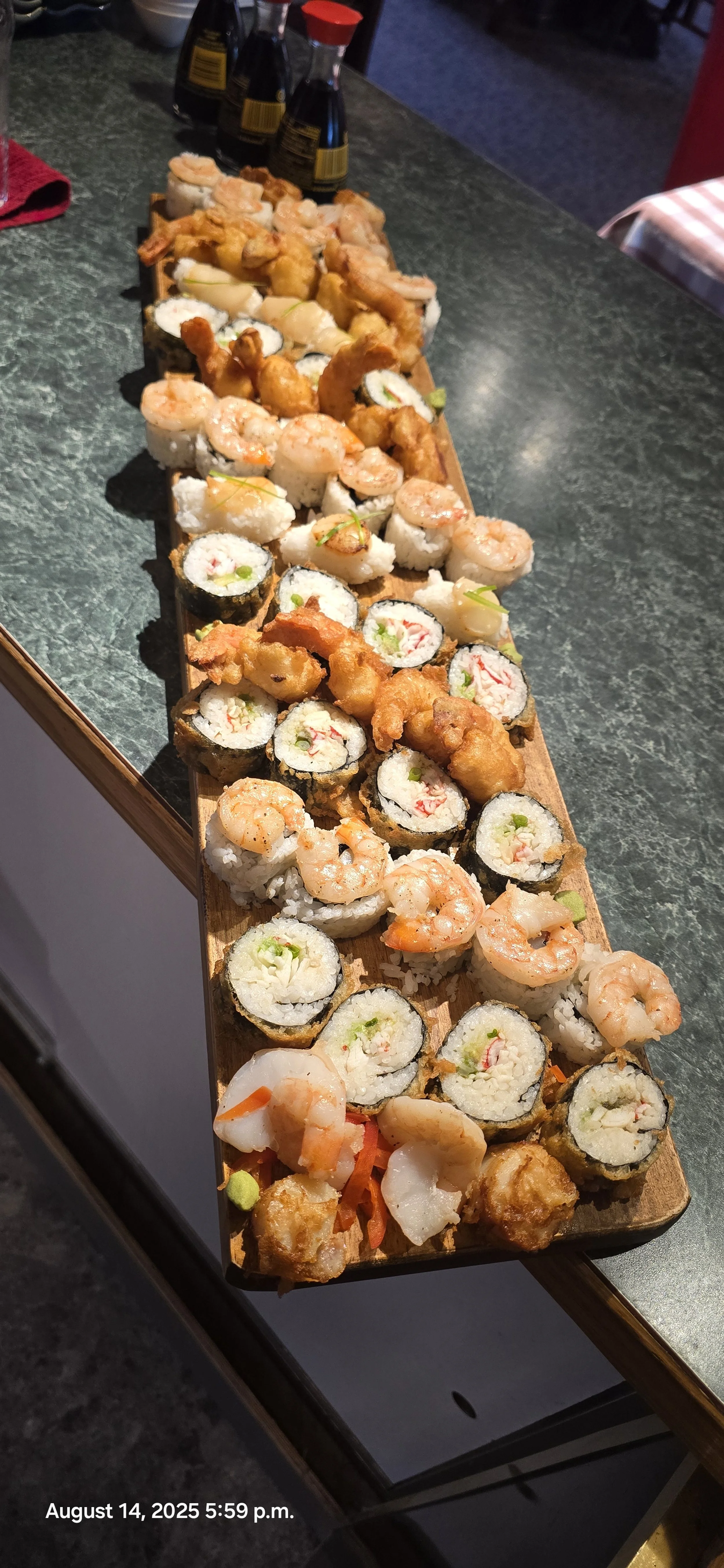 A long wooden tray filled with assorted sushi rolls, including shrimp, fish, and vegetable varieties. There are soy sauce bottles and condiments at the back of the table.