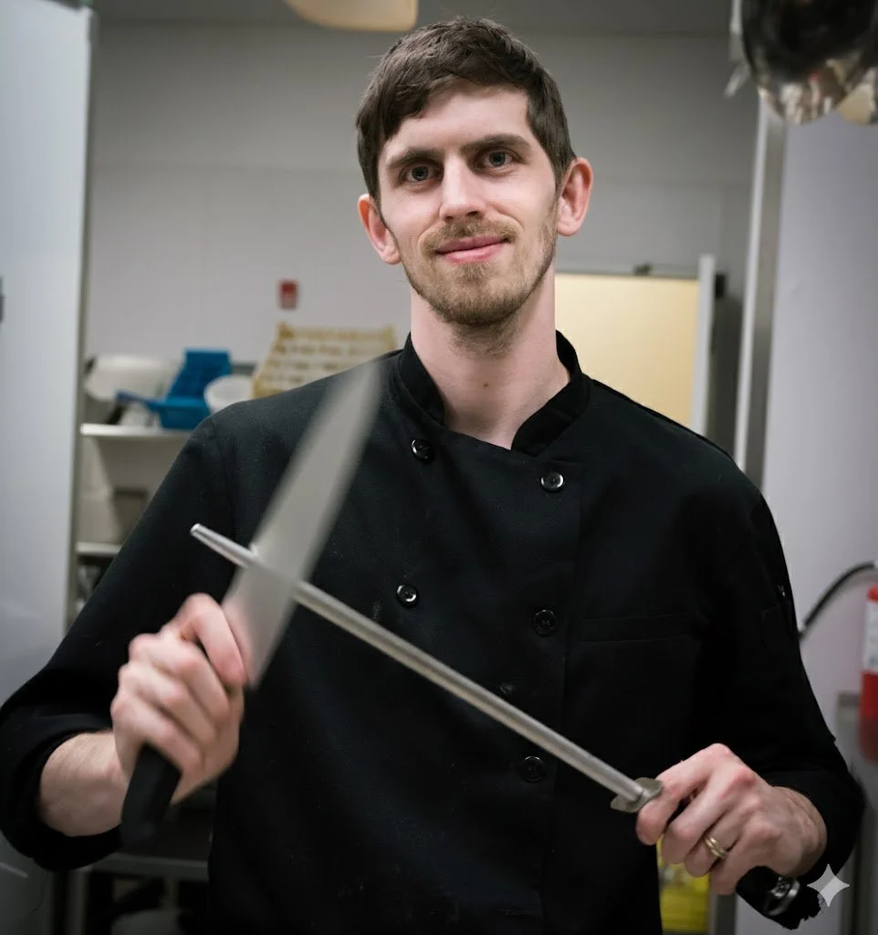 A young man in a black chef's coat holding a large kitchen knife in a professional kitchen.