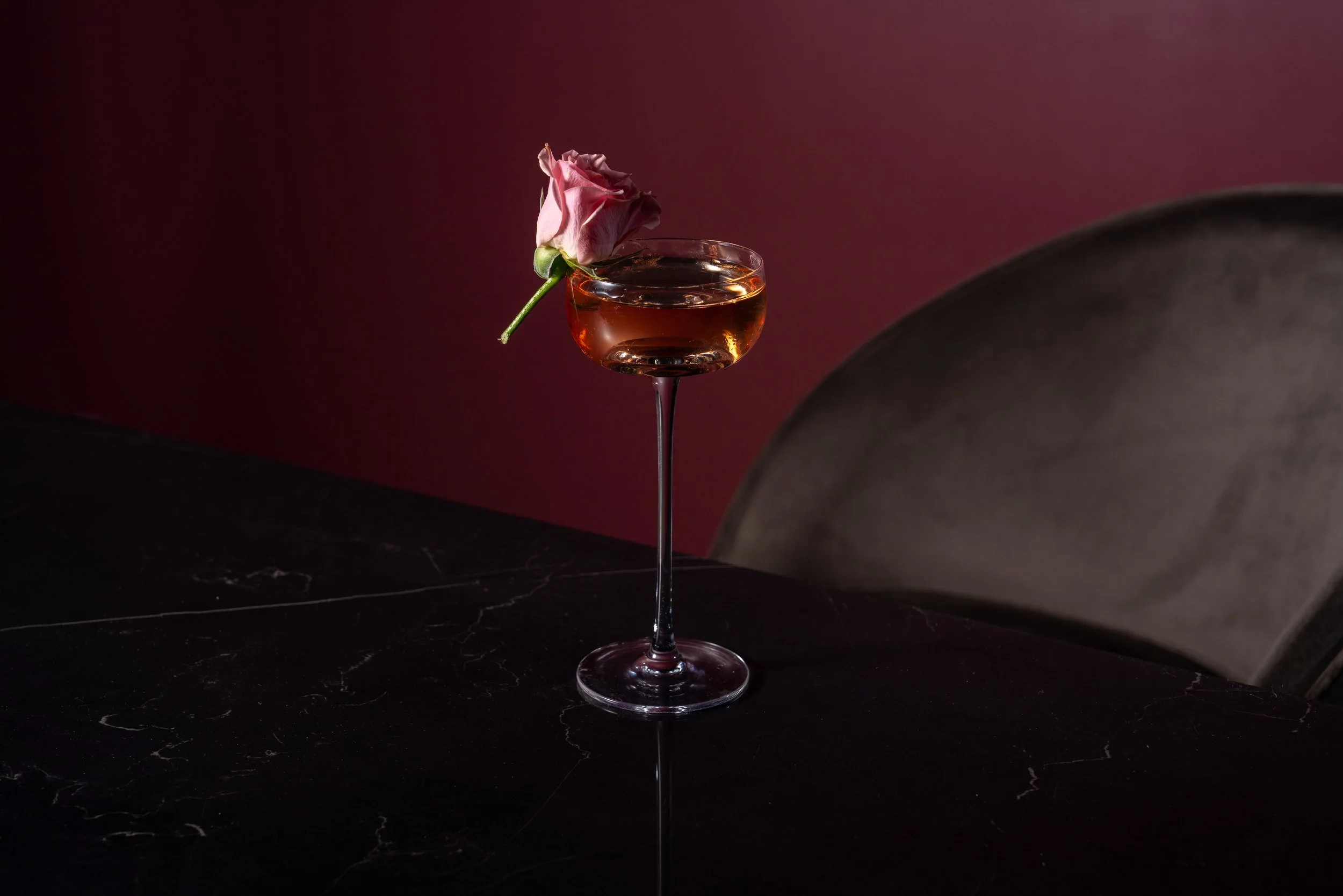 Image of a cocktail glass with a long stem topped with a pink rose on the rim