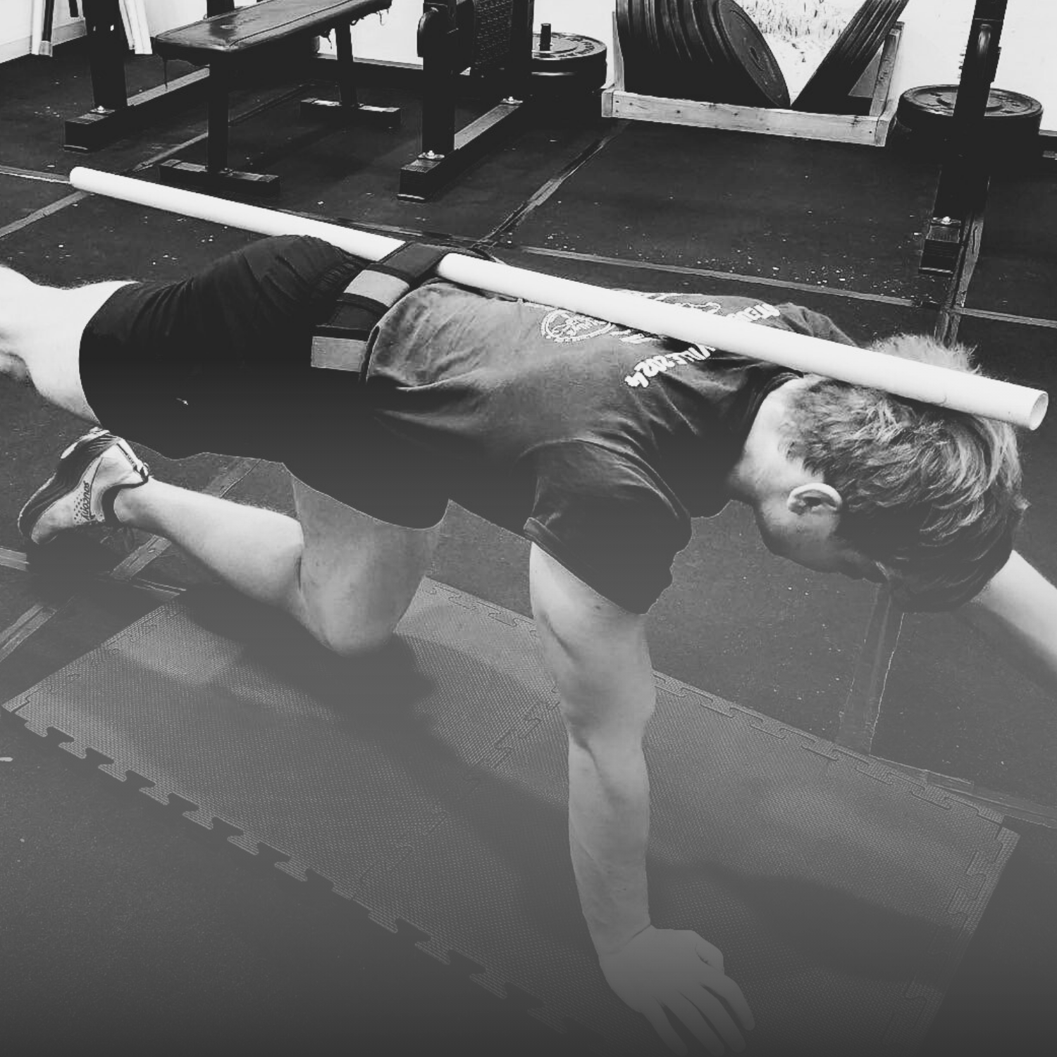 A person doing a mountain climber exercise on a gym mat with a PVC pipe resting on their back, in a gym setting.