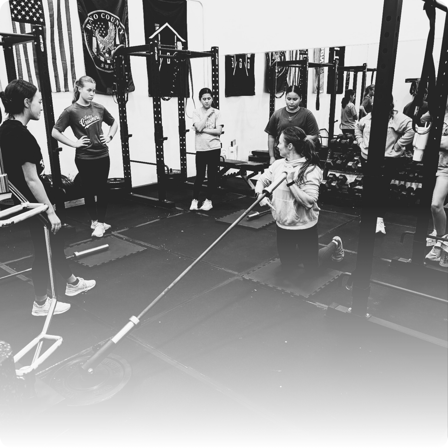 Women in a gym participating in a workout class, with one woman kneeling while using a barbell, and others standing around watching.
