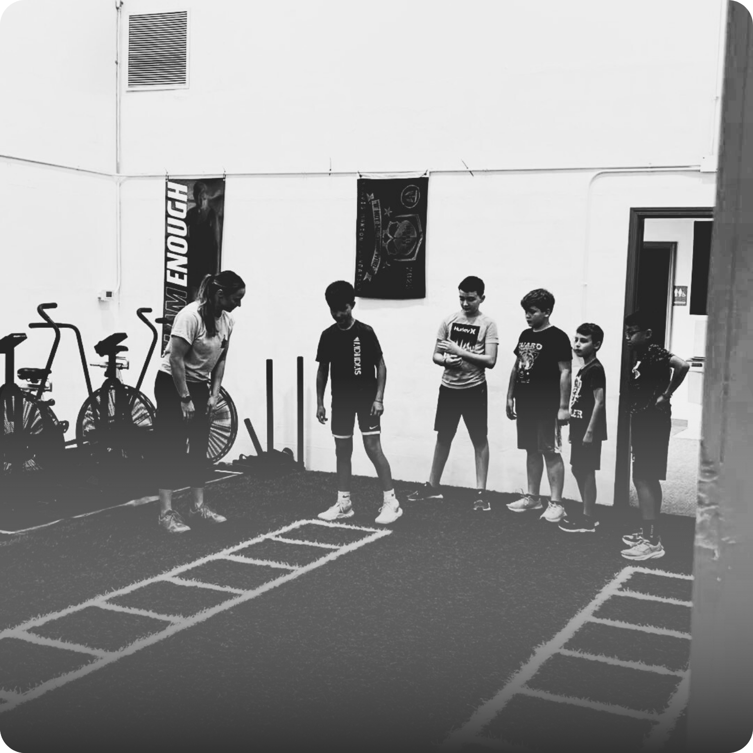 Group of children and a woman in a gym, standing in line, with exercise equipment and banners in the background.