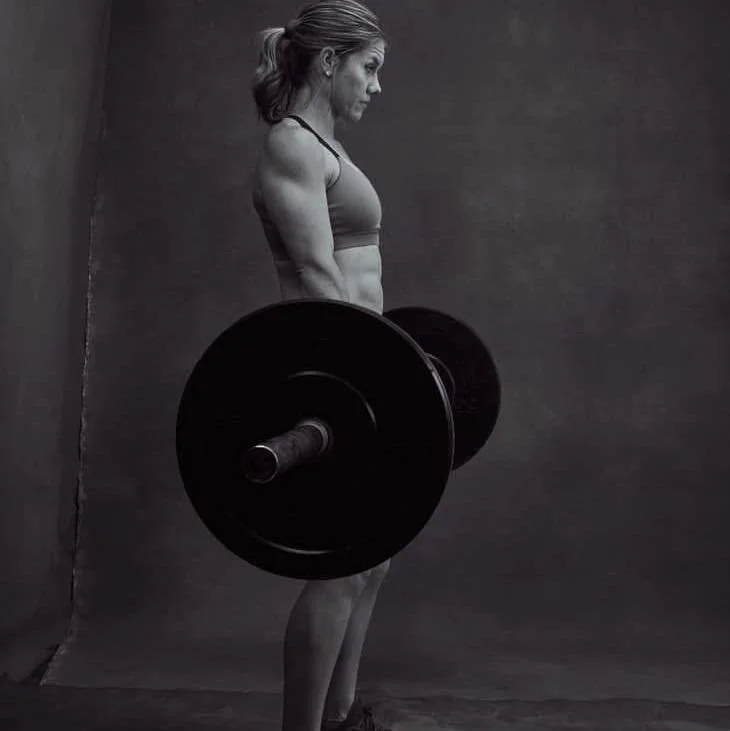 Black and white photo of a woman with toned muscles lifting a barbell, standing against a plain background.