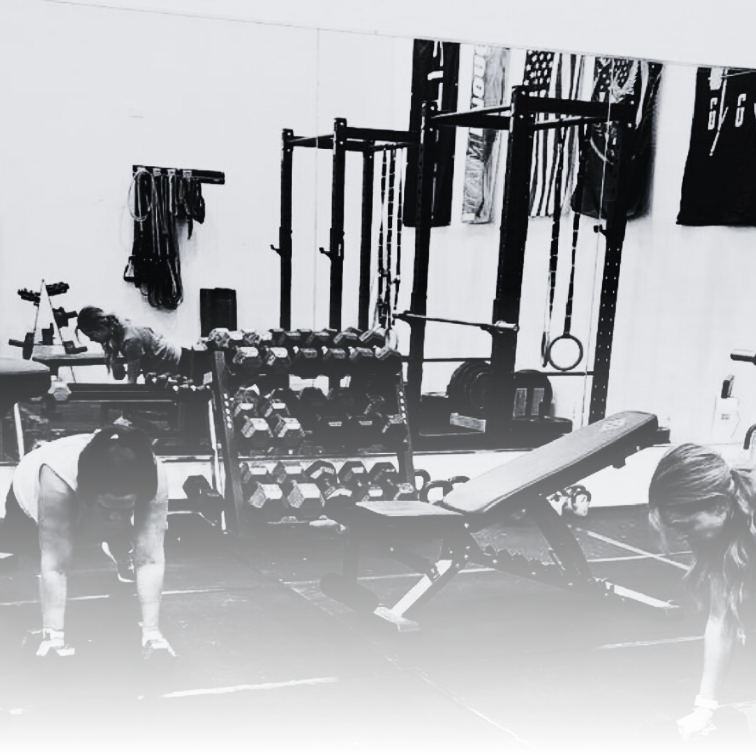 Two women are stretching in a gym with exercise equipment including dumbbells, a weight rack, and a workout bench.