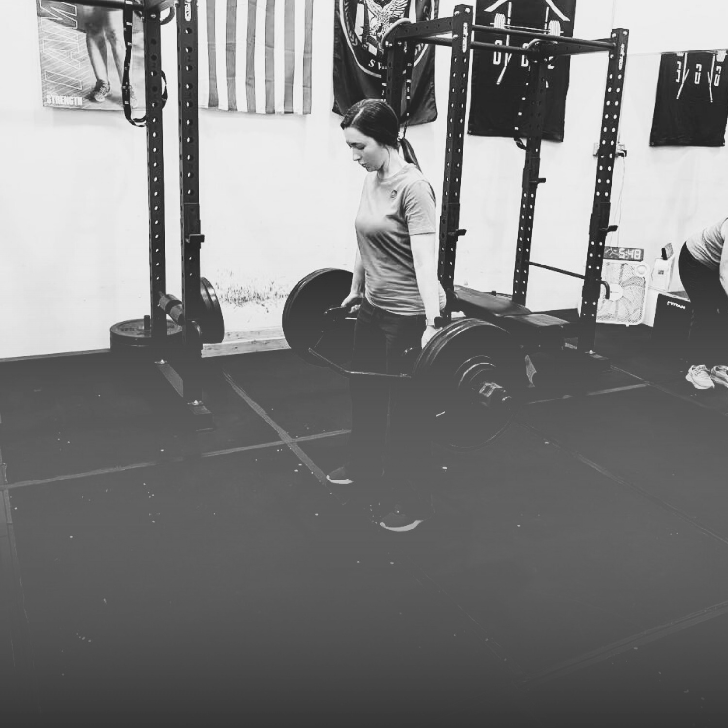 A woman in a gym lifting a heavy barbell with weights in a black-and-white photo. The gym has banners hanging on the wall and a small fan in the background.