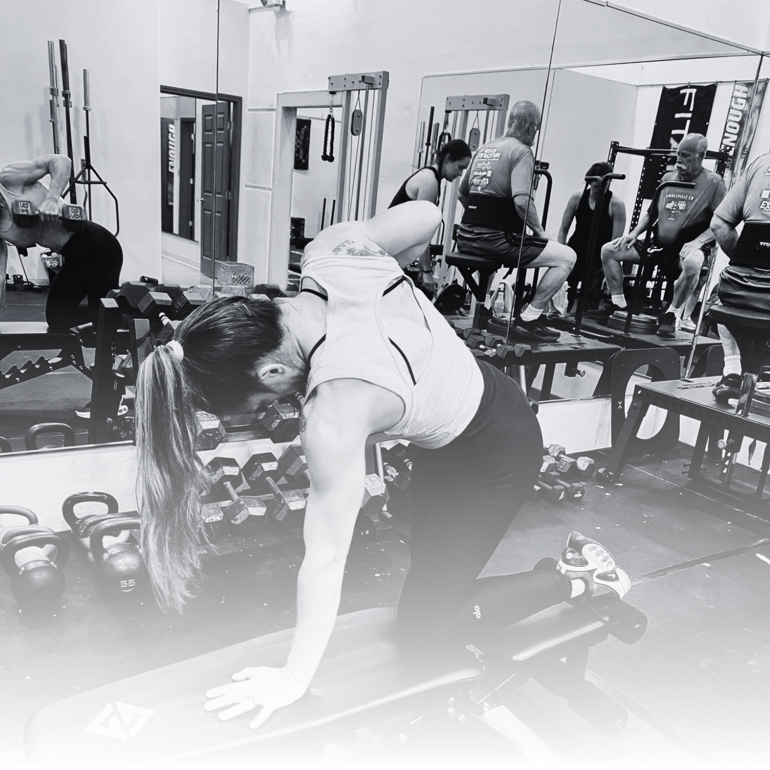 A woman with long hair in a ponytail doing a workout on a bench in a gym, with weights and exercise equipment visible around her, and other people exercising in the background.